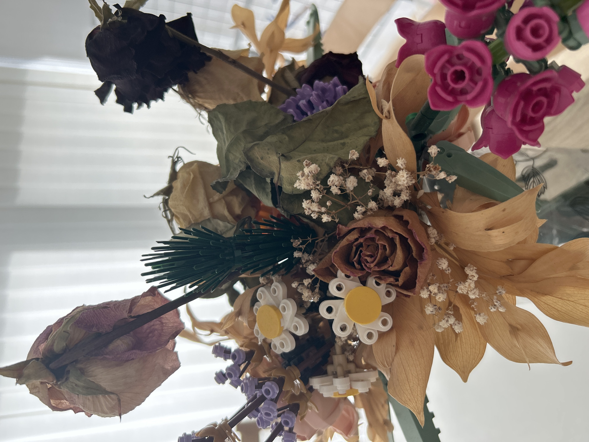A close-up, overhead shot shows a bouquet of dried flowers and LEGO flower bricks arranged in a clear vase on a reflective surface, likely a table. The dried flowers exhibit various shades of brown, beige, and pink, with some showing signs of wilting and decay. Notable are a large dried pink rose at the top left, a dark brown dried rose at the top right, and several smaller dried roses and baby's breath throughout the arrangement. Interspersed with the dried flora are LEGO bricks, forming white daisy-like flowers with yellow centers, purple flower elements, and green leaf-like pieces. A cluster of vibrant pink LEGO roses is visible in the lower right corner. The background consists of a softly blurred white curtain or blinds, suggesting an indoor setting with natural light. The scene is static, with no people or discernible action. The image does not provide any information about its location in Varna, Bulgaria.