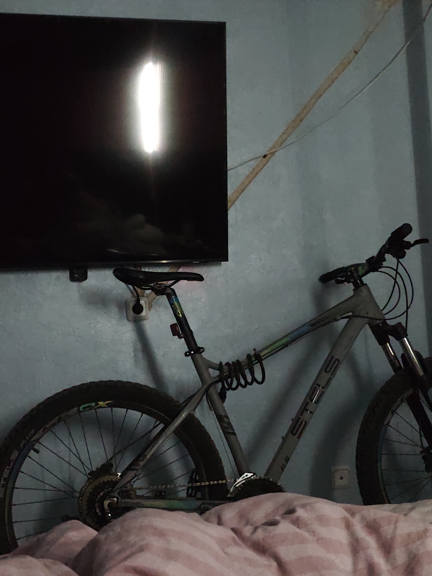 The image displays an indoor scene featuring a grey full-suspension mountain bicycle leaning against a light blue wall. A black flat-screen television is mounted on the wall above the bicycle, with a bright, vertical light reflection visible on its screen. A white electrical outlet is positioned on the wall behind the bicycle's seat. A light-colored cable extends diagonally upwards on the wall, passing behind the TV.
The bicycle's frame prominently features the brand name "STELS" in large letters. The rear wheel tire also has "STELS PLUS" and "CX" visible on its side. A black coiled bicycle lock is attached to the bike's frame. In the immediate foreground, part of a bed covered with a pinkish, patterned duvet or blanket is visible, suggesting the room is a bedroom or a living space.
No people are present in the image, and no specific activity is taking place. The time of day is not clearly discernible from the indoor lighting, although the bright reflection on the TV screen indicates a light source in the room. Weather conditions cannot be determined. The provided location context is Chaikovsky, Russia, though there are no visual cues within the image to confirm this. No particular emotions are conveyed.