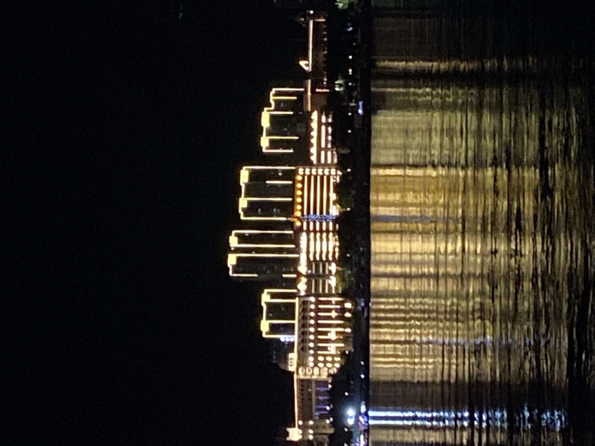 The image shows a nighttime view of a waterfront cityscape with illuminated buildings. The architecture includes several tall, modern structures with bright yellow accent lighting, as well as a longer, more ornate building with decorative lighting. The buildings are reflected in the dark water in the foreground, creating shimmering patterns of light. The sky is black, indicating it is nighttime. The scene is located in Blagoveshchensk, Russia, on the Amur River, which forms part of the border between Russia and China.