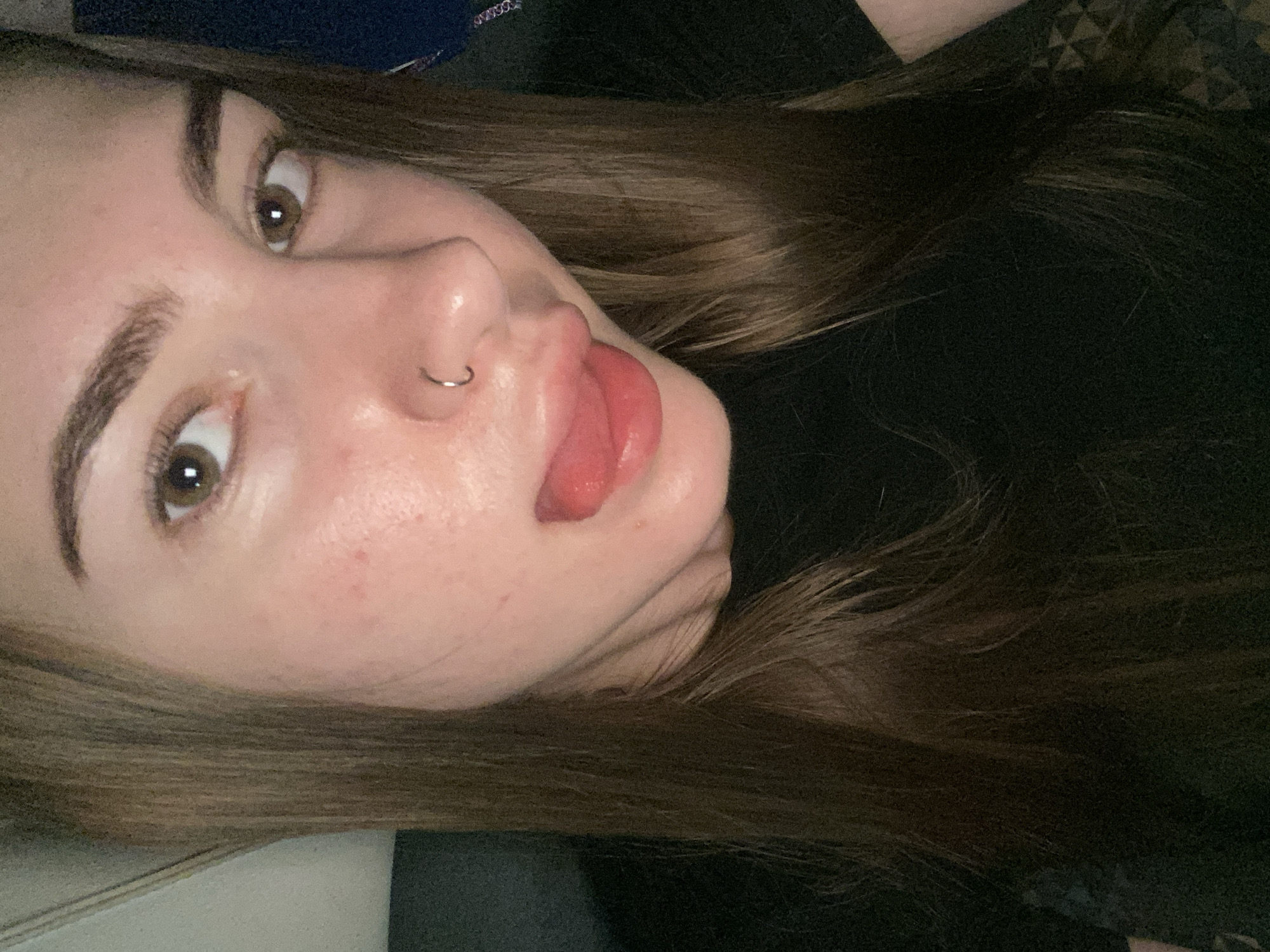 A close-up, forward-facing, headshot of a young Caucasian woman with long, brown hair. She is wearing a black shirt and has a silver hoop piercing in her nose. Her lips are slightly parted and her tongue is visible. The background is dark, with some texture visible on the right. The lighting is dim and appears to be coming from the front. The image quality is fair, with some noise and a slight blur. There are no discernible landmarks or environmental details that would suggest a specific location within Дьяково, Russia.