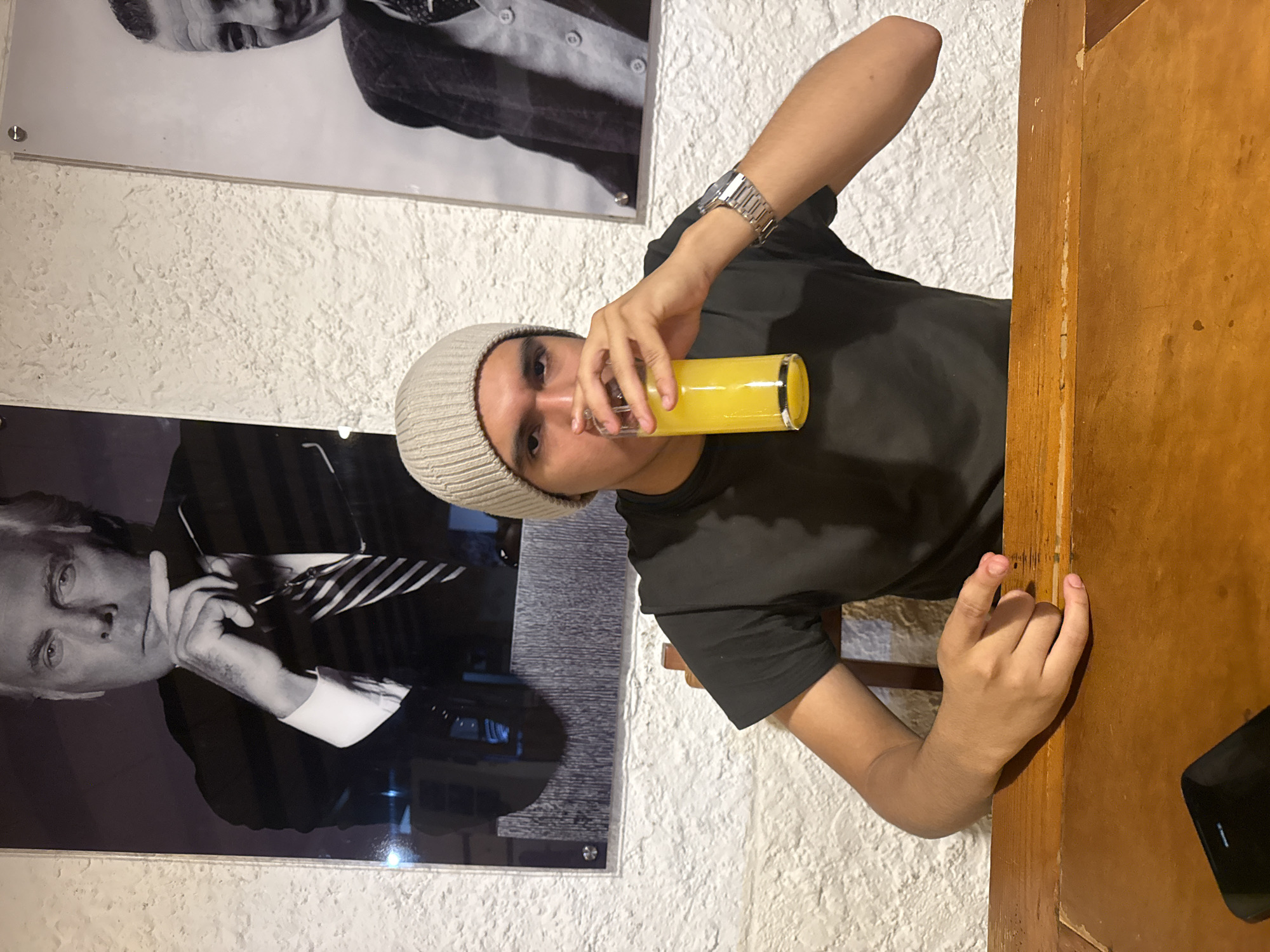 A medium shot depicts a young man drinking from a glass of orange juice. He is wearing a grey beanie and a dark t-shirt, and a silver watch is visible on his left wrist. He is sitting at a wooden table, and part of a black smartphone is on the table, near the right edge of the frame. Behind him, two framed black-and-white portraits are hanging on a white, textured wall. The location is presumed to be Chipilo de Francisco Javier Mina (Chipilo), Mexico.
