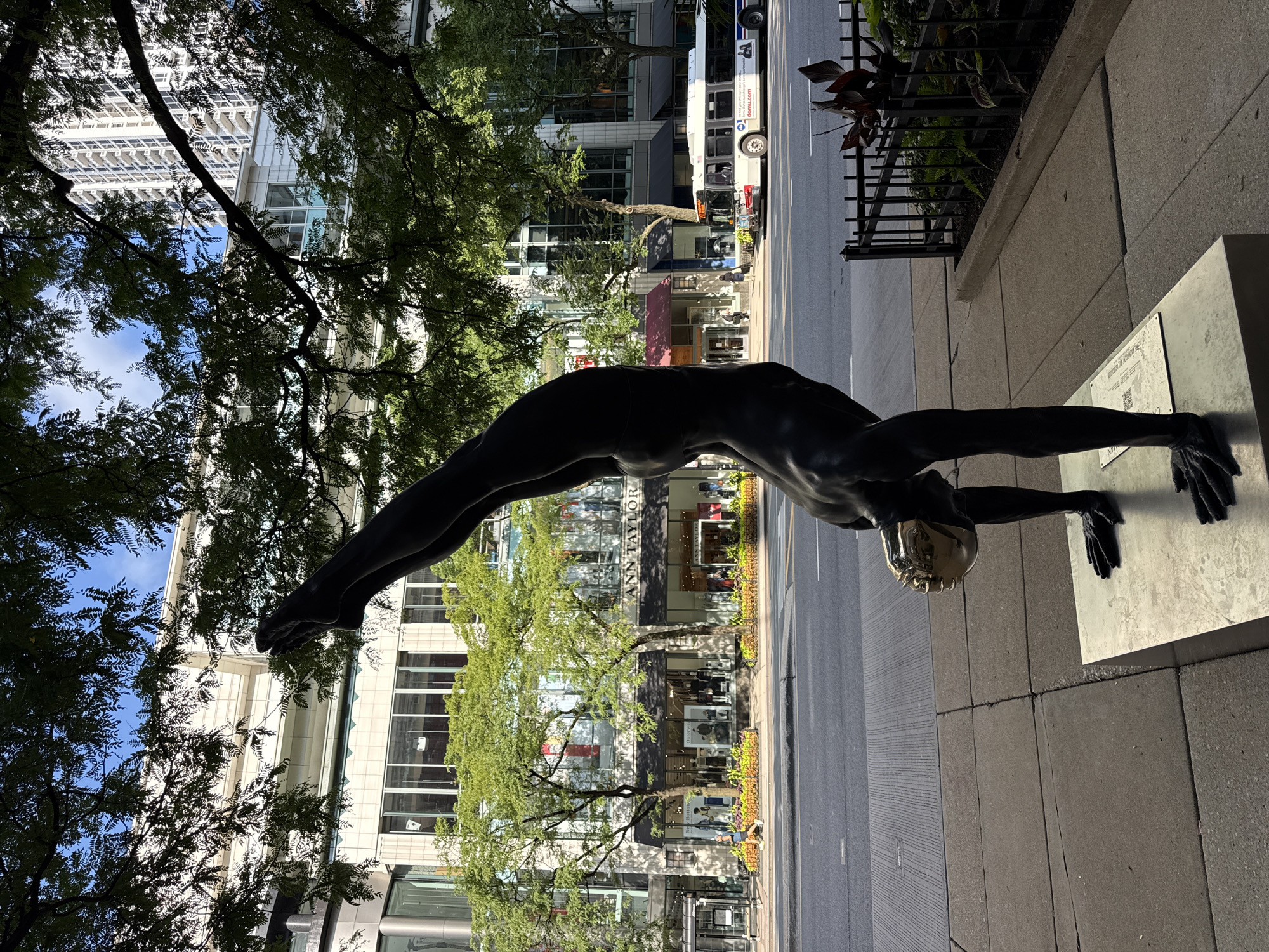 A full-body dark bronze sculpture of a male gymnast performing a handstand is positioned on a polished metal pedestal on a sidewalk in Chicago, USA. The gymnast's body is in a vertical line, supported by his hands, with his legs extended upwards. His head is covered by a mirrored gold-colored mask that reflects the sky. The sculpture is situated in front of a row of shops, including "Ann Taylor," and a tall apartment building is visible in the background. A street with a bus and traffic runs parallel to the sidewalk, separated by a curb. Trees with green foliage cast shadows over the scene, and a black metal fence encloses a small planter area to the right of the sculpture. The sky is blue with scattered white clouds.