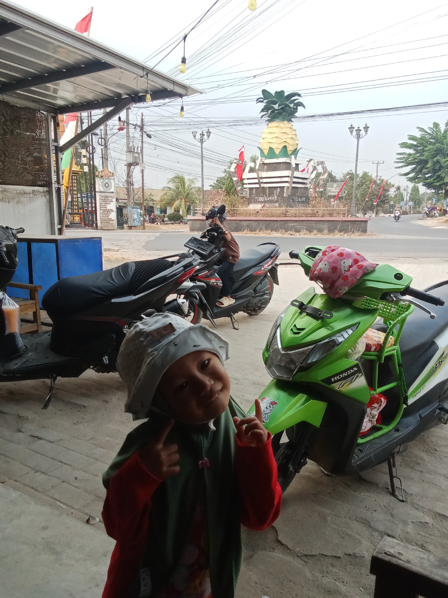 A child wearing a light-colored hat and a red shirt with a green jacket is standing in front of two parked motorcycles. The child is looking towards the camera and has their fingers pointed outwards from their face. The motorcycles are a black Honda and a green Honda. In the background, there is a large pineapple monument labeled "TUGU NANAS" which is a landmark in Toto Katon, Indonesia. There are also several power lines, streetlights, and some buildings visible in the distance. The scene appears to be outdoors, possibly on a street or near a local establishment.