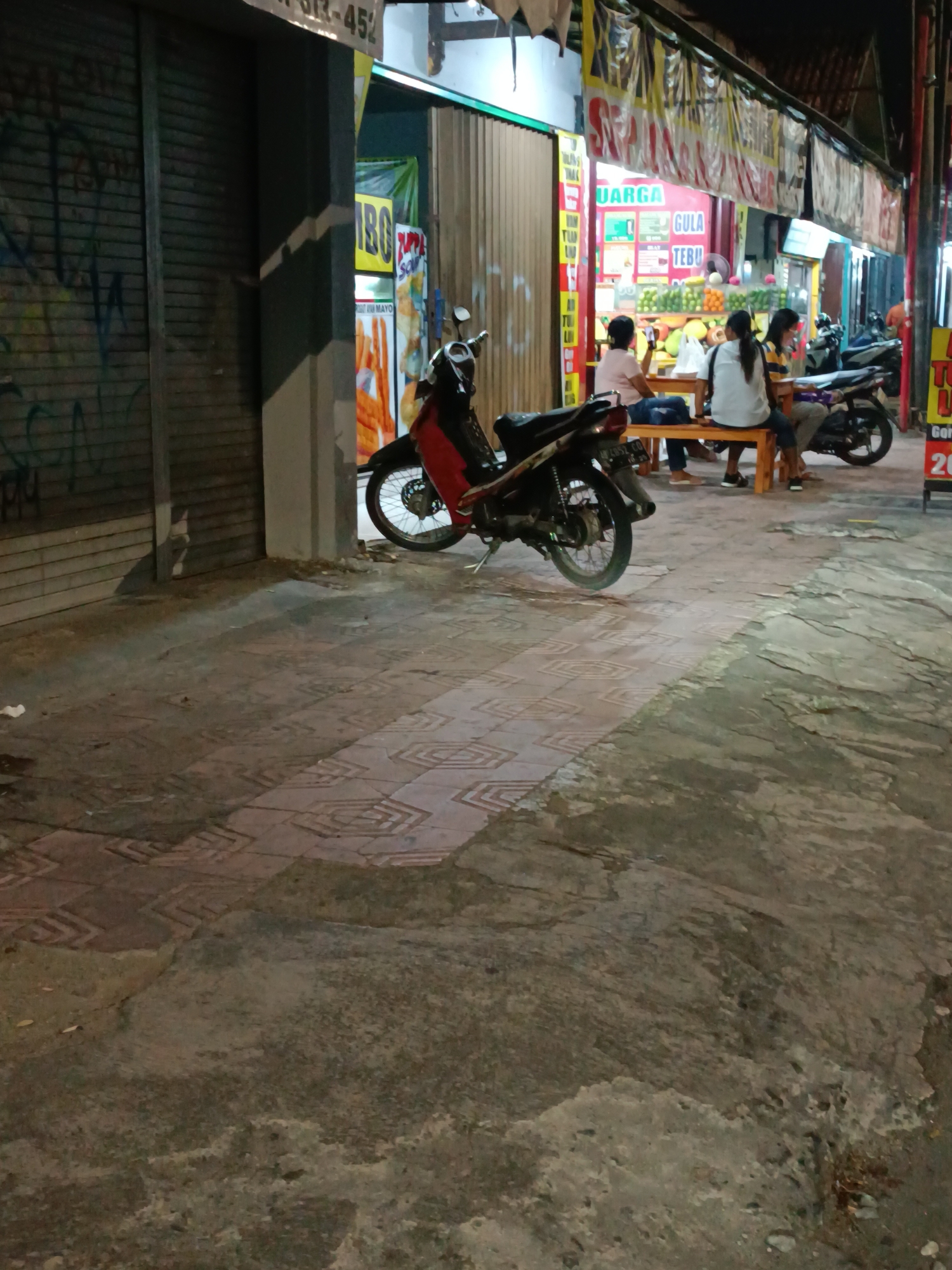 The image depicts a nighttime scene on a street in Yogyakarta, Indonesia. A red and black motorcycle is parked on the patterned sidewalk. Behind the motorcycle, a row of shops is visible, some with their metal shutters down. One open shop displays a variety of food and drinks, with a banner overhead featuring Indonesian text. Three people are seated at a wooden table in front of this shop, appearing to be customers. The ground is a mix of concrete and patterned tiles, with some debris scattered around. The overall atmosphere suggests a commercial area at night, possibly a food stall or small restaurant.