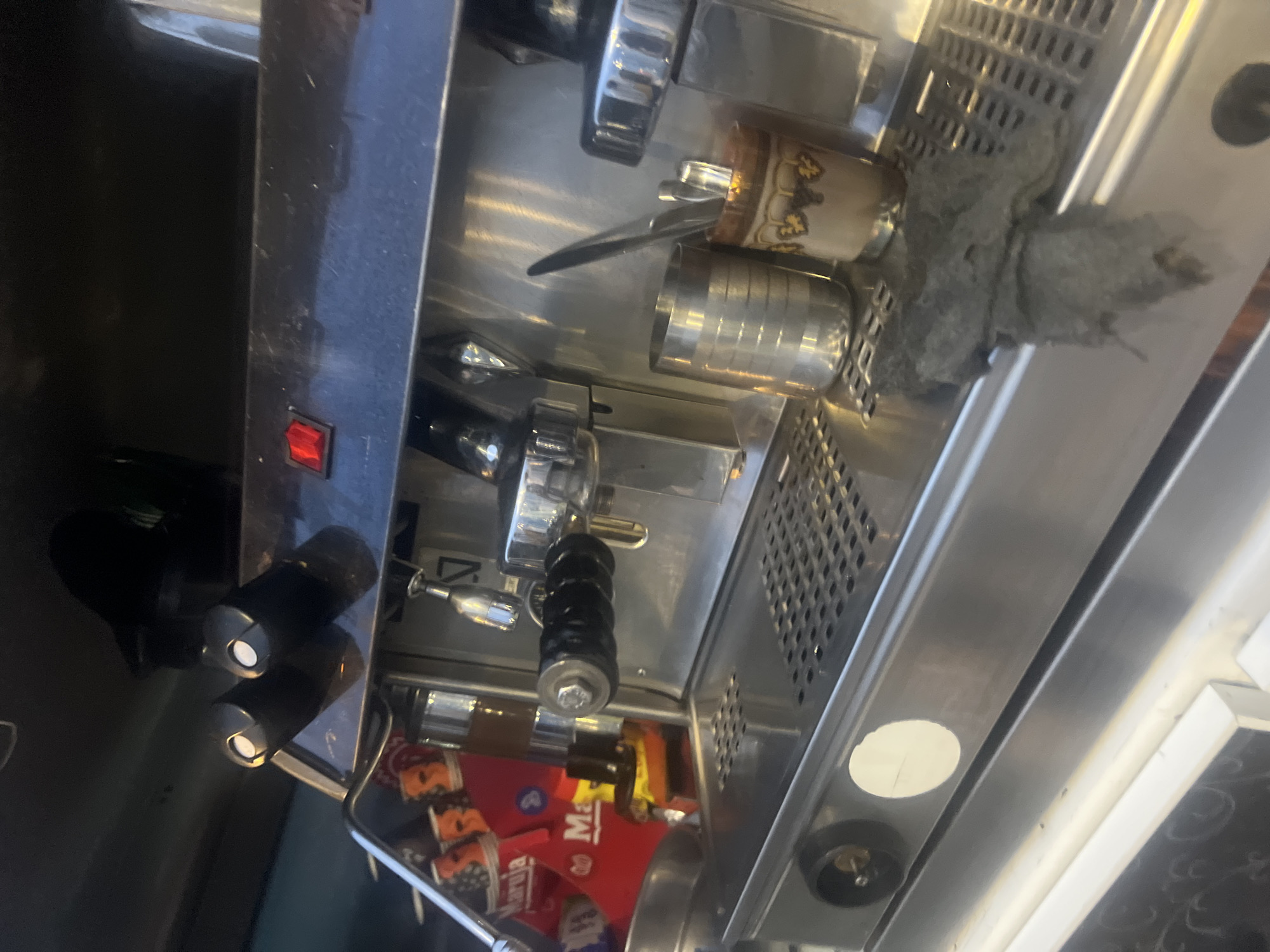 The image shows a close-up of an espresso machine, possibly located in Martil, Morocco, given the presence of "Martil" in a graphic below the machine. The machine is stainless steel and appears to be in use, with a towel partially covering the top. Coffee cups are situated on top. The machine has several controls and dispensers. The power switch is visible on the side.