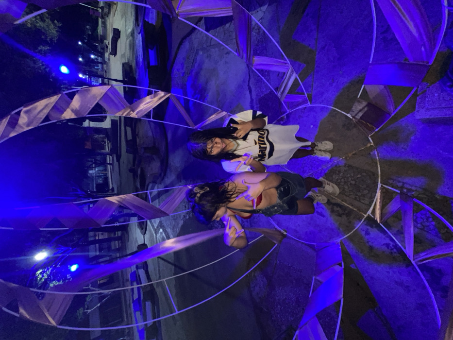 Two women stand inside a circular structure with fabric and blue lighting. One woman wears a white t-shirt and glasses, making a peace sign with her left hand. The other wears a red top and denim shorts, also making a peace sign. The structure casts shadows and reflections, creating an interesting perspective. The photo is taken from above, looking down on the women and the geometric shapes of the structure. A building and street lights are visible in the background.