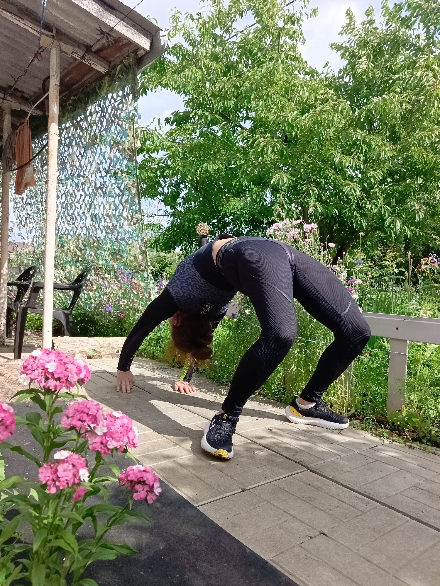 A person is doing a backbend on a paved pathway outdoors, in what appears to be a backyard or garden area. The person is wearing black athletic leggings with a textured pattern, and a long-sleeved dark shirt with a lighter pattern. Their feet are flat on the ground, with knees bent and hands on the pathway behind them, supporting their arched back. The environment is lush and green with trees and flowering plants visible. A camouflage net hangs from a wooden structure to the left, and two black plastic chairs are partially visible. Pink flowers are in the foreground. The location is identified as Vyazovka, Russia.