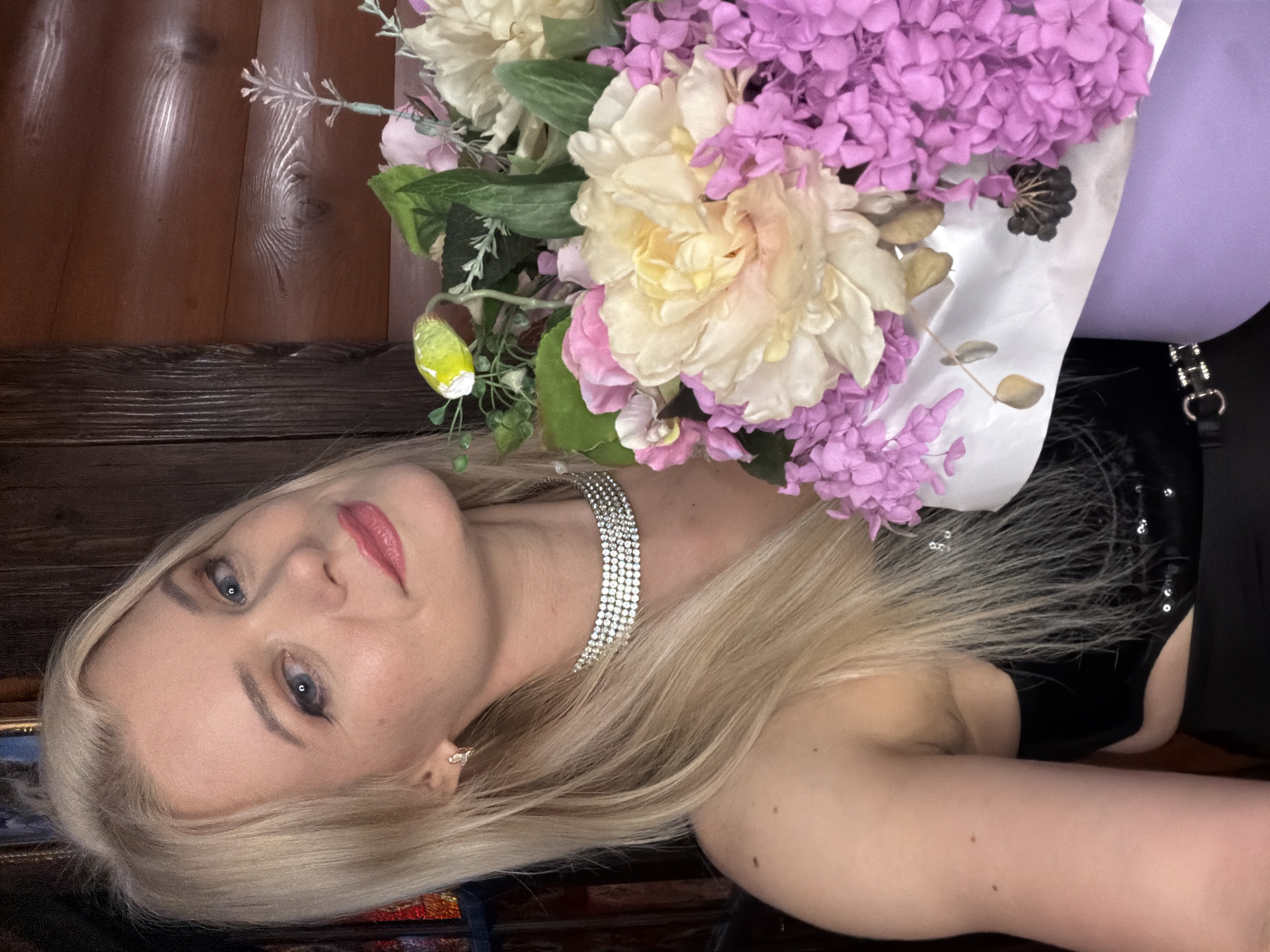An adult Caucasian female with blonde hair and blue eyes is pictured in a close-up shot. She is wearing a black, strappy top adorned with sequins and a sparkling choker necklace. The woman is holding a bouquet of large, pale yellow and pink flowers, interspersed with smaller purple blossoms. The background appears to be a wooden interior, possibly a room with wood-paneled walls. The overall impression is that of a portrait taken indoors, perhaps at a private event or residence in Sochi, Russia, given the context of the inquiry.