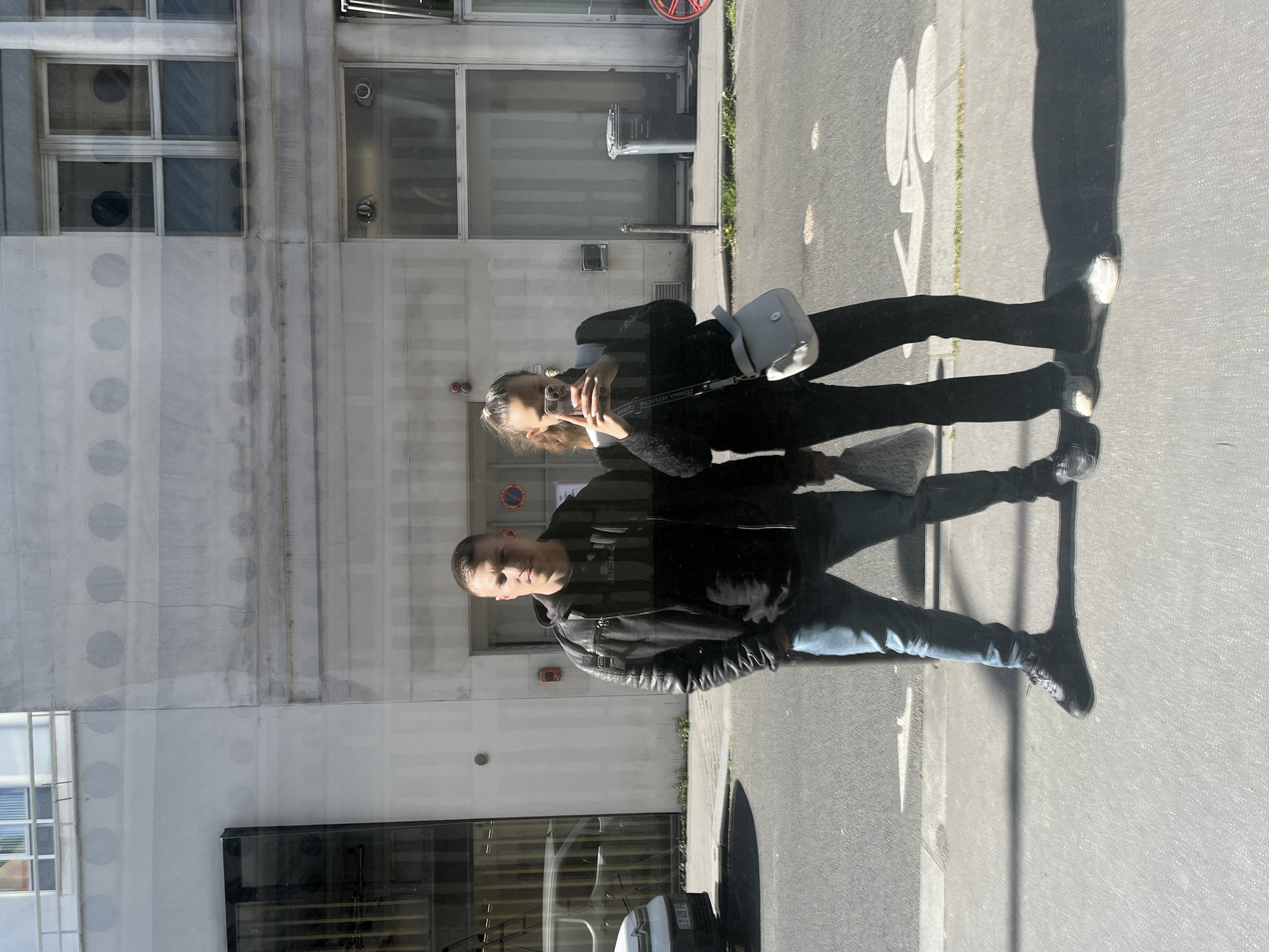 The image displays two individuals, a man and a woman, captured as reflections in the large glass facade of a modern urban building. The scene takes place outdoors on a bright, sunny day, evidenced by distinct, elongated shadows cast on the asphalt road and sidewalk.
The female subject, positioned towards the left of the frame, is dressed in dark attire and carries a light-colored, possibly white, cross-body bag. She holds a smartphone, appearing to be taking a photograph or video, likely a selfie or of the male subject beside her, through the reflective surface. The male subject, to her right, is wearing a dark leather jacket over a dark shirt and blue jeans. Both individuals appear to be standing still, facing the glass building.
The building itself is a light-colored structure, possibly concrete, featuring vertical lines and some internal or reflected circular patterns. The street includes a sidewalk, an asphalt road, and a partially visible white painted bicycle symbol on the ground.
Notable details include the strong sunlight, indicating a clear-sky day. The female subject's handbag strap clearly shows the brand name "TOMMY HILFIGER" in reverse due to the reflection. A reflection of a white van is visible near the male subject's lower body, with part of its license plate, "125 FH," discernable. The layered nature of the reflection superimposes the people and street environment onto the building's structure, creating a dynamic visual effect. The urban setting and visible cues align with the given location context of Paris, France.