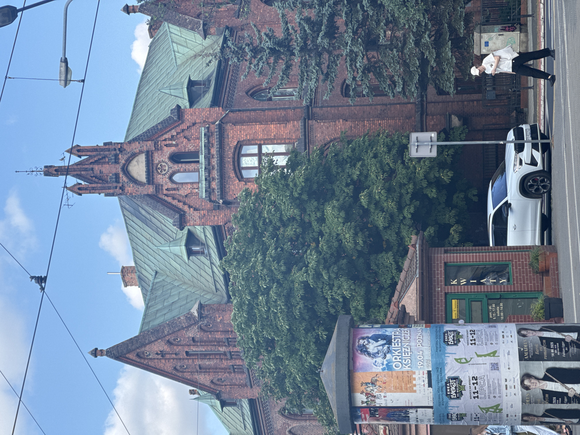 The image depicts a street scene in Poznan, Poland, with a prominent brick building featuring a green tiled roof and Gothic architectural elements. A large tree with lush green foliage partially obscures the building, while evergreen trees are visible on the right. In the foreground, a cylindrical advertisement pillar displays various posters, including one for "ORKIESTRA KSIĘŻNICZEK 19.04 POZNAŃ". To the right of the pillar is a small kiosk with a green door and the word "KWIATY" (flowers) written vertically on the facade. A white SUV is parked on the street, and a person wearing a white cap, t-shirt, and dark pants is walking across the street. Overhead, power lines crisscross the sky under a partly cloudy blue sky. The overall impression is a sunny day in an urban environment, likely in the center of Poznan, given the architectural style and the presence of public advertisements.