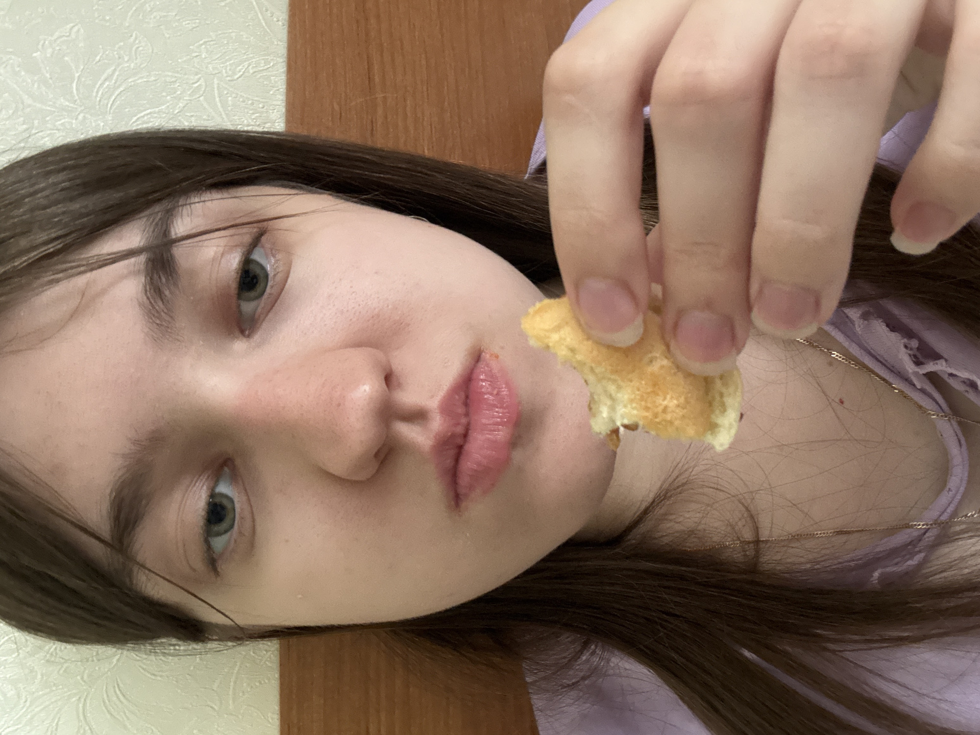 A close-up portrait of a young woman holding a partially eaten piece of golden-brown food in front of her mouth. She has fair skin, dark brown hair, and light green eyes. She is wearing a light purple top. A gold necklace is visible on her neck. The background includes a light-colored wall with a faint pattern and a vertical wooden panel. The location is Unknown, Unknown.