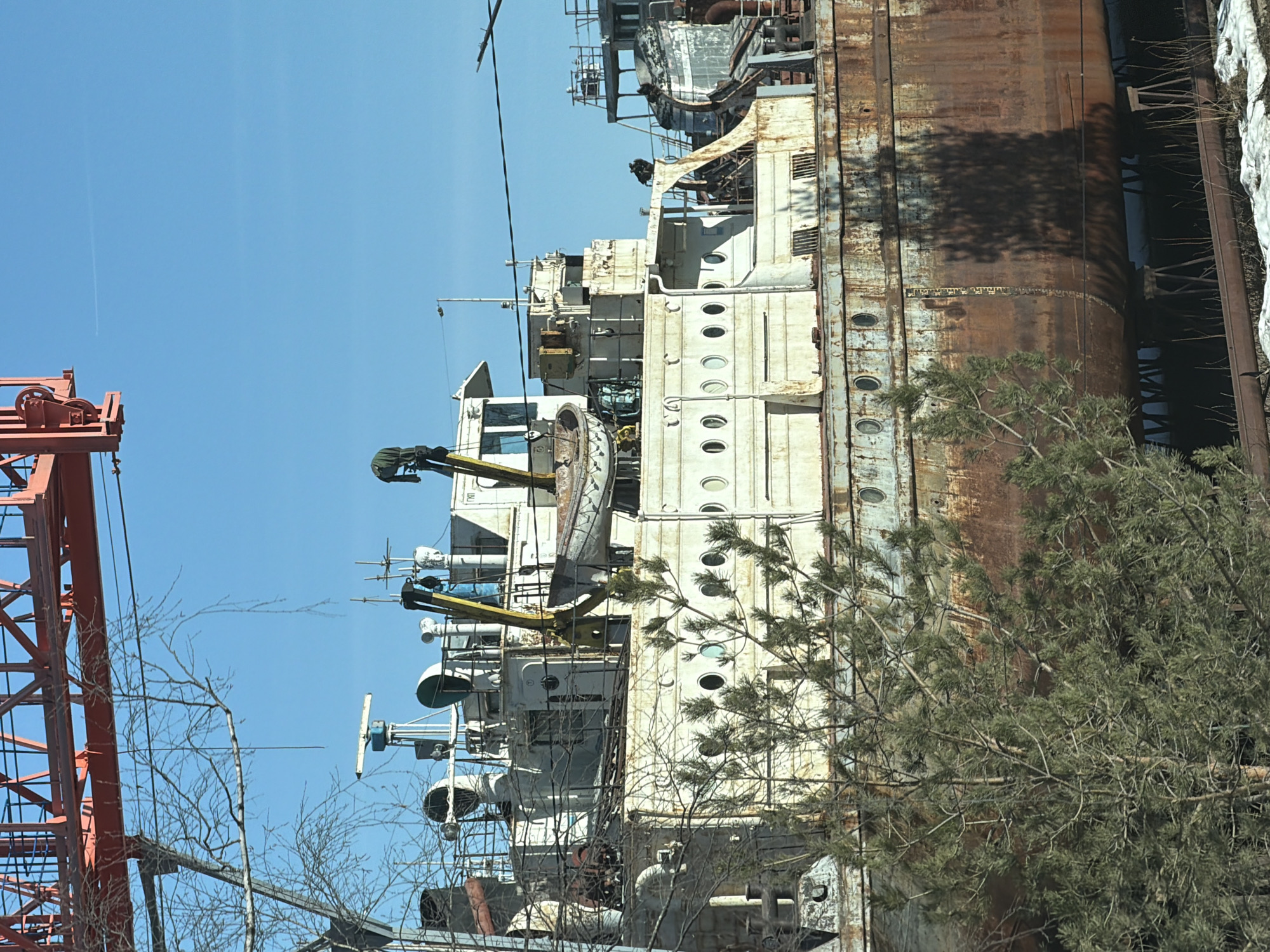 An image of a decaying, partially dismantled ship hull or superstructure. The visible section features multiple rows of porthole-style openings on white-painted sections, adjacent to heavily rusted vertical plating. A tree with bare branches in the foreground partially obscures the view. Sections of a red crane are visible on the left side of the frame. The image is oriented vertically, which distorts the perspective of the ship. This is most likely a ship located in the city of Chaikovsky, Russia, based on prior details.
