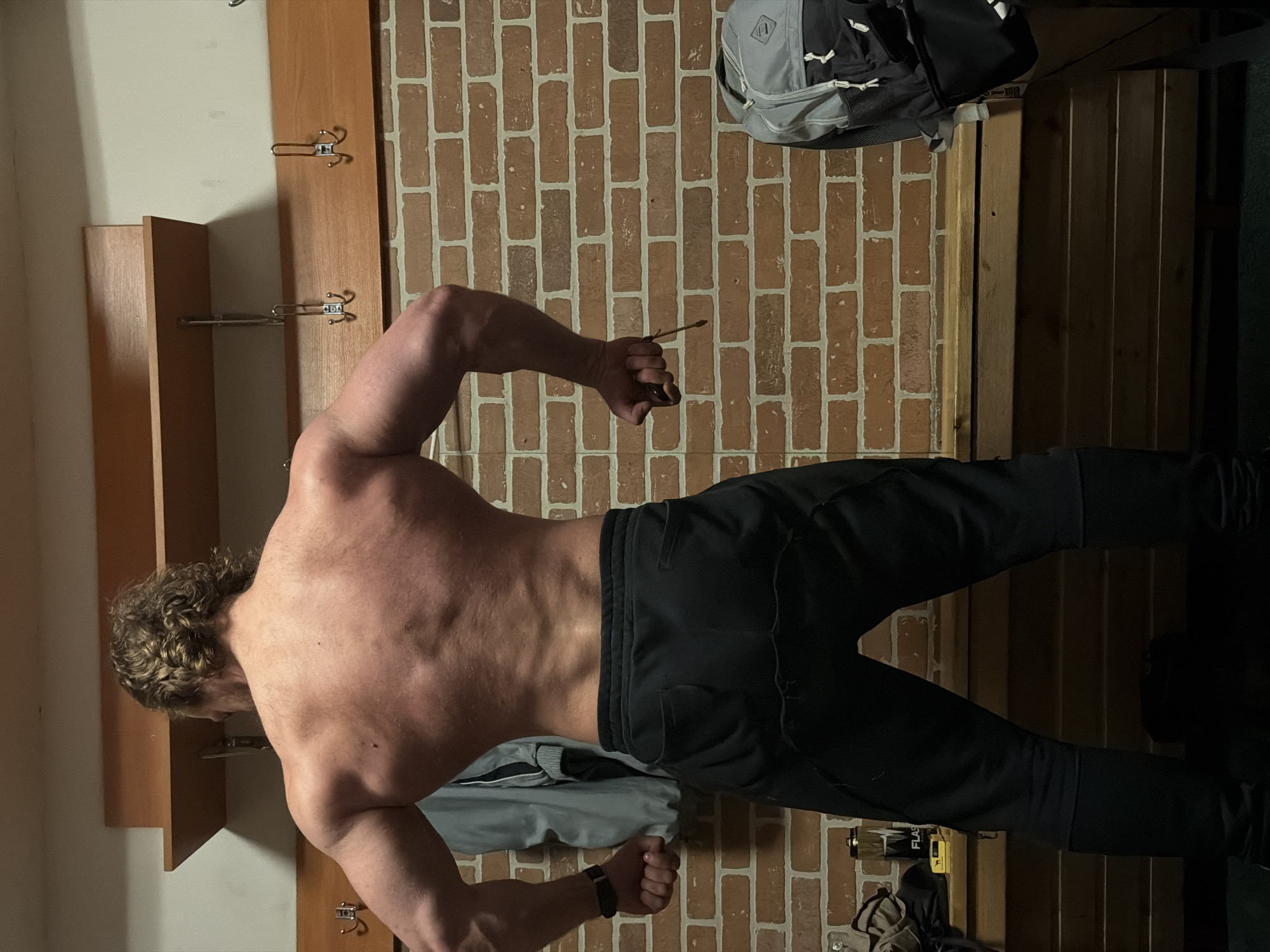 The image displays a male individual, photographed from the back, standing in what appears to be a changing room or locker room. The individual is shirtless, exhibiting a developed musculature in the back, shoulders, and arms. His dark, curly hair reaches the upper back. He is wearing black athletic pants. His arms are flexed outwards with clenched fists; a dark fitness tracker is visible on his left wrist, and his right hand holds a small, dark object with a light-colored string.
The background consists of a reddish-brown brick wall. Mounted on the wall are wooden fixtures: a narrow shelf positioned high up, a wider horizontal wooden panel below it featuring multiple metal double-pronged coat hooks, and a wooden slatted bench along the lower portion of the wall.
To the left, a light grey jacket with black detailing hangs from a hook. On the bench beneath it, a black and silver water bottle branded "FLASH" with a lightning bolt logo is visible next to a small yellow object. To the right, a grey and black backpack with multiple compartments is situated on the bench or floor.
The environment is illuminated by artificial light. The floor is dark and mostly obscured. The precise location within Saint Petersburg, Russia, cannot be determined from the visual information provided. The setting is consistent with a communal changing area typically found in a gymnasium or sports complex.