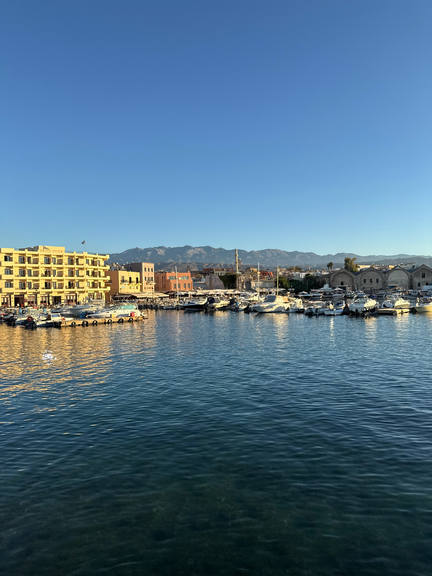 In an aerial, eye-level shot, a bright blue sky stretches over a harbor filled with moored boats. The sun illuminates the water, creating a shimmering reflection of a yellow building on the left side of the frame. The building has multiple floors and numerous windows, and a flag flies from its roof. Behind the boats, a collection of buildings in various colors and architectural styles line the waterfront, with mountains visible in the distance. The water itself is a deep blue, with small ripples on its surface.