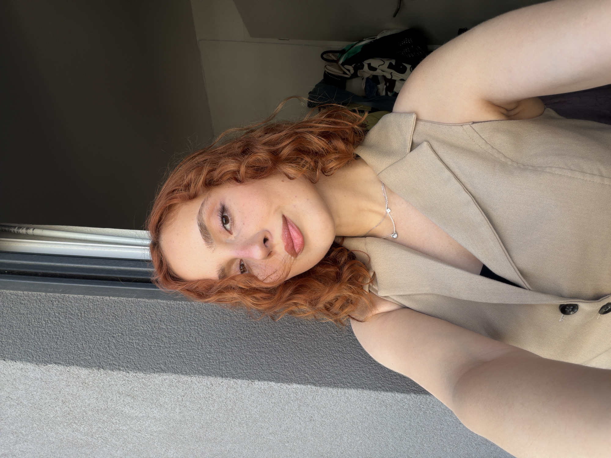 A young woman with reddish-orange curly hair is posing for a selfie. She is wearing a light beige sleeveless vest over a dark top and a delicate silver necklace. Her makeup includes eyeshadow and blush. The background shows a textured grey wall on the left and a dark interior space on the right with some hanging items. The lighting suggests it is daytime, with sunlight casting shadows. The image does not contain any specific landmarks or context that would indicate a location in the city of Strasbourg, France.