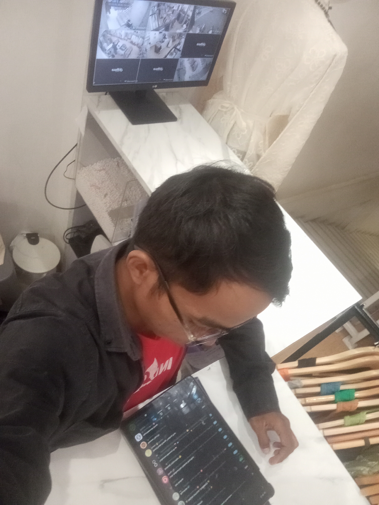 A man is seated at a white table, looking down at a tablet. He is wearing a dark jacket over a red shirt and glasses. To his left is a computer monitor displaying multiple security camera feeds. Behind the man, to the right, is a white draped object, possibly a mannequin covered with a cloth. In the background, shelves are visible containing various wooden items, some wrapped with colored thread or ribbon. The scene appears to be indoors, possibly an office or a retail space, in the city of Larangan, Indonesia.