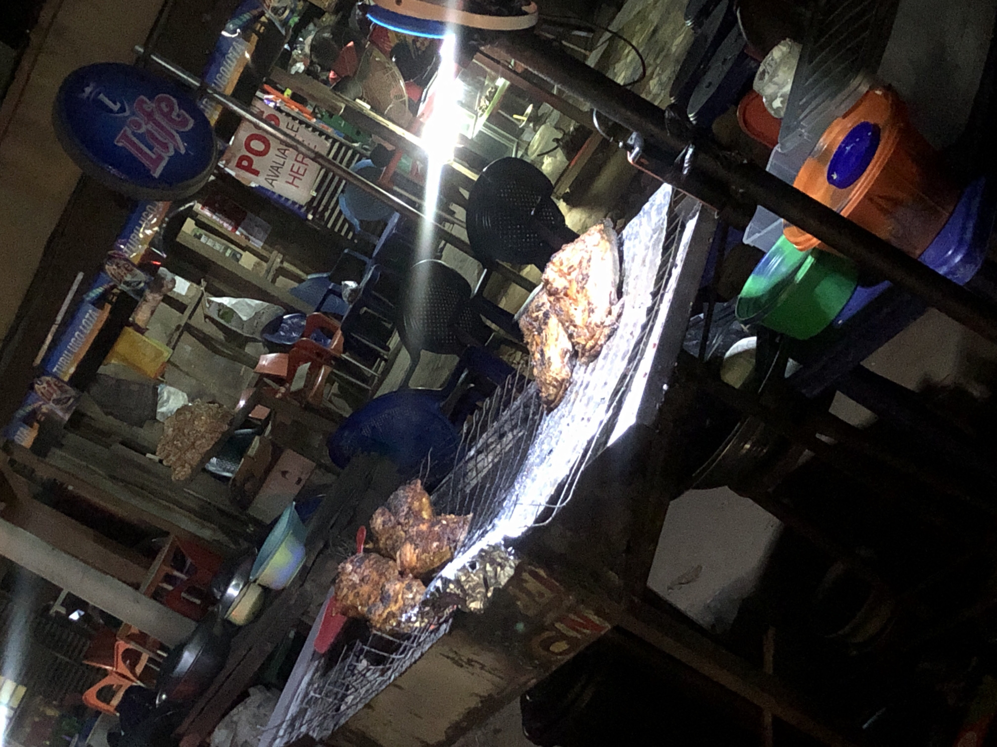 This image shows a vibrant street scene in Onitsha, Nigeria at night. A grill with what appears to be chicken is front and center. An advertisement for "Life" beer is visible. There are an abundance of various containers and chairs. The scene is lit with harsh artificial light. There are no people visible in the frame. The environment is dense and cluttered.