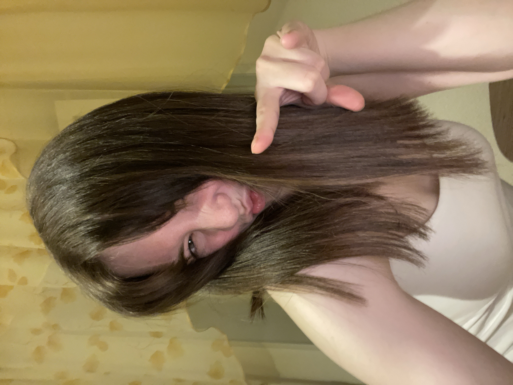 A person with long, dark brown hair, light skin, and wearing a white top is making a funny face at the camera. They are pointing a finger down towards their hair. The background appears to be an interior space with a light yellow wall. There is no readily available information to confirm that this photo was taken in Chaikovsky, Russia.