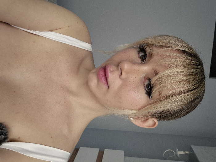 The image displays a close-up, head-and-shoulders portrait of a female individual in an indoor setting. The subject has a light skin tone with visible freckles distributed across her face, neck, and upper chest. Her hair is blonde with darker roots, styled with a fringe (bangs) over her forehead and appears to have a crimped or wavy texture, with longer strands pulled back. Her eyes are dark brown, accentuated with dark eyeliner and long eyelashes. Her lips are colored with a pink lipstick. She wears a white or light-colored garment with visible shoulder straps. A small, light-colored stud earring is present in her right earlobe. She maintains direct eye contact with the camera, with her head slightly tilted.
The background consists of a plain light grey wall. To the right of the subject's head, a white wall-mounted light fixture, designed to resemble a candle sconce with a curved arm, is visible. Below the light fixture, a section of white furniture featuring a horizontal brown accent line is partially visible. A dark, fluffy object is present at the bottom right edge of the frame. The visual evidence within the image does not provide specific identifiers to confirm the location as Batumi, Georgia.