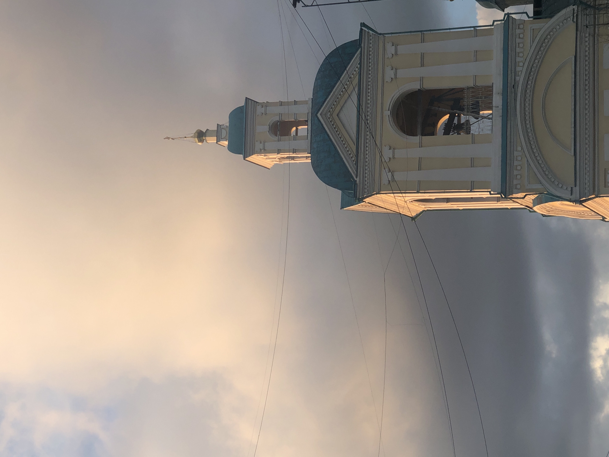 A church bell tower is seen from a low angle against a sky of varying shades of gray. The tower is a cream color with turquoise roofing and a gold spire. It features arched windows and decorative architectural details. Power lines crisscross the scene. This appears to be a church in Krasnoyarsk, Russia.