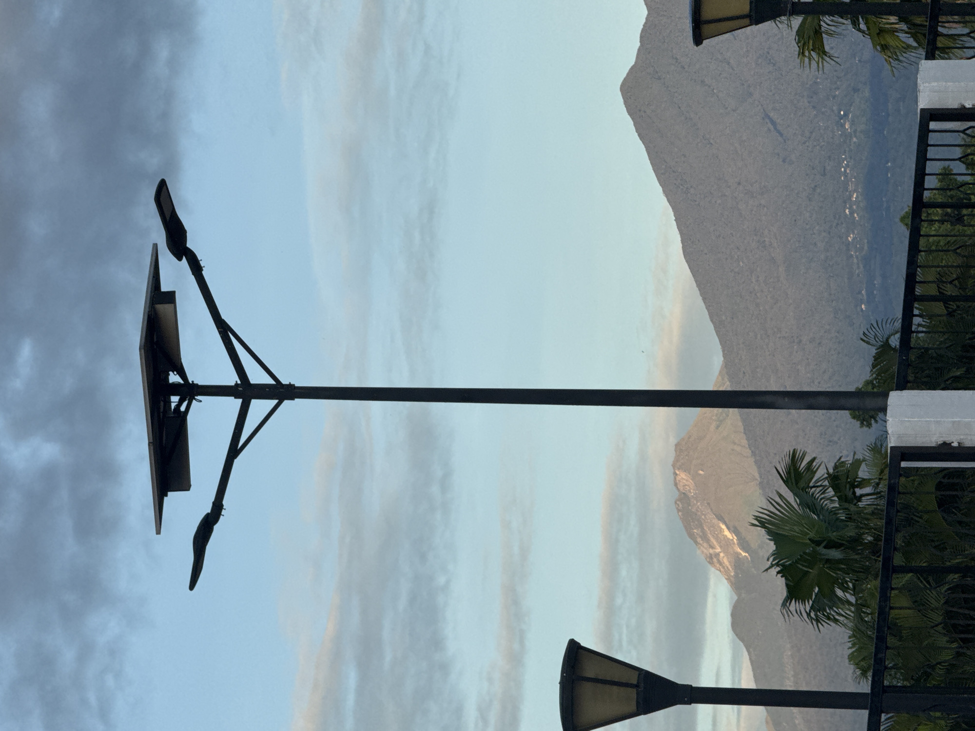 The image shows a street scene in Davao City, Philippines, at what appears to be dusk or dawn. The foreground contains two black metal light poles and a solar panel mounted on one. The light poles are typical street lamps with curved hoods. The sky is light blue with streaks of grey clouds. In the background is a mountain, partially illuminated by the sun. A black metal fence and some palm trees are also visible. The scene lacks people, but the presence of street lamps and a solar panel suggests a built environment. The background is dominated by the mountain, indicative of the geography of the region.