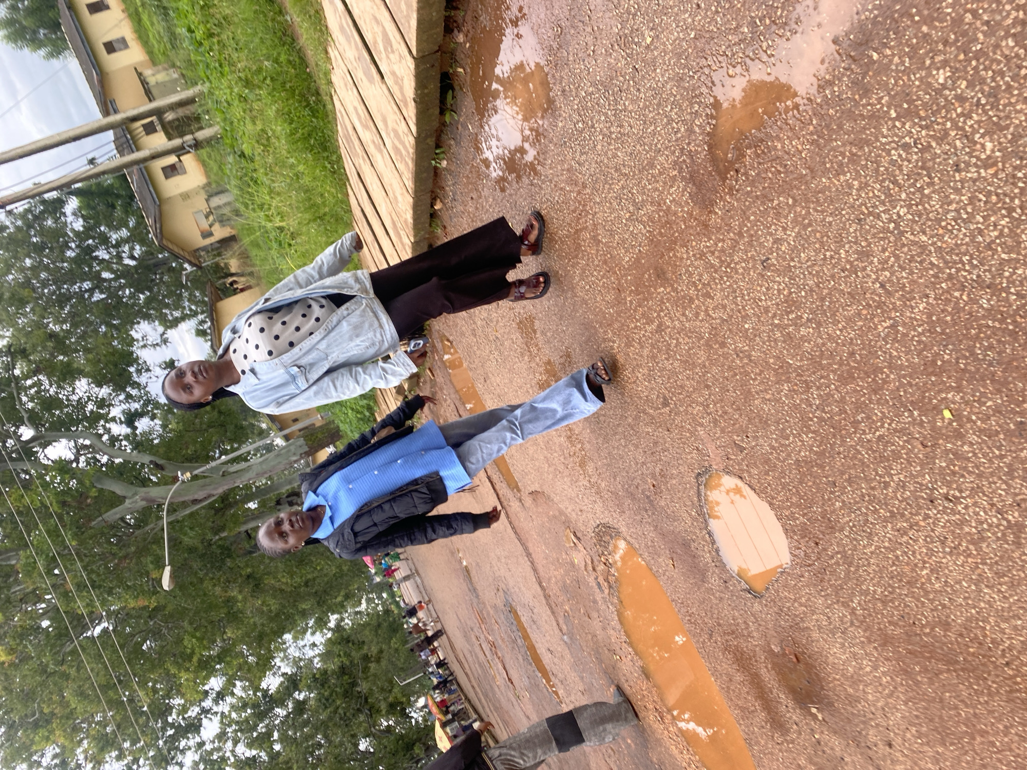 The image depicts a street scene in Rafin Par, Nigeria, characterized by a heavily potholed and muddy road, indicative of recent rainfall. Two young women are walking on the road. The woman on the left is wearing a black jacket over a blue collared shirt and light-colored pants. The woman on the right is wearing a light blue denim jacket over a white and black polka dot shirt and dark pants. In the background, several small buildings with yellow walls and corrugated iron roofs are visible, along with lush green vegetation and trees. Power lines run across the upper part of the image. Several market stalls with colorful umbrellas are also present further down the street, suggesting a commercial area. The overall environment appears to be a typical Nigerian town or city street after a period of rain.