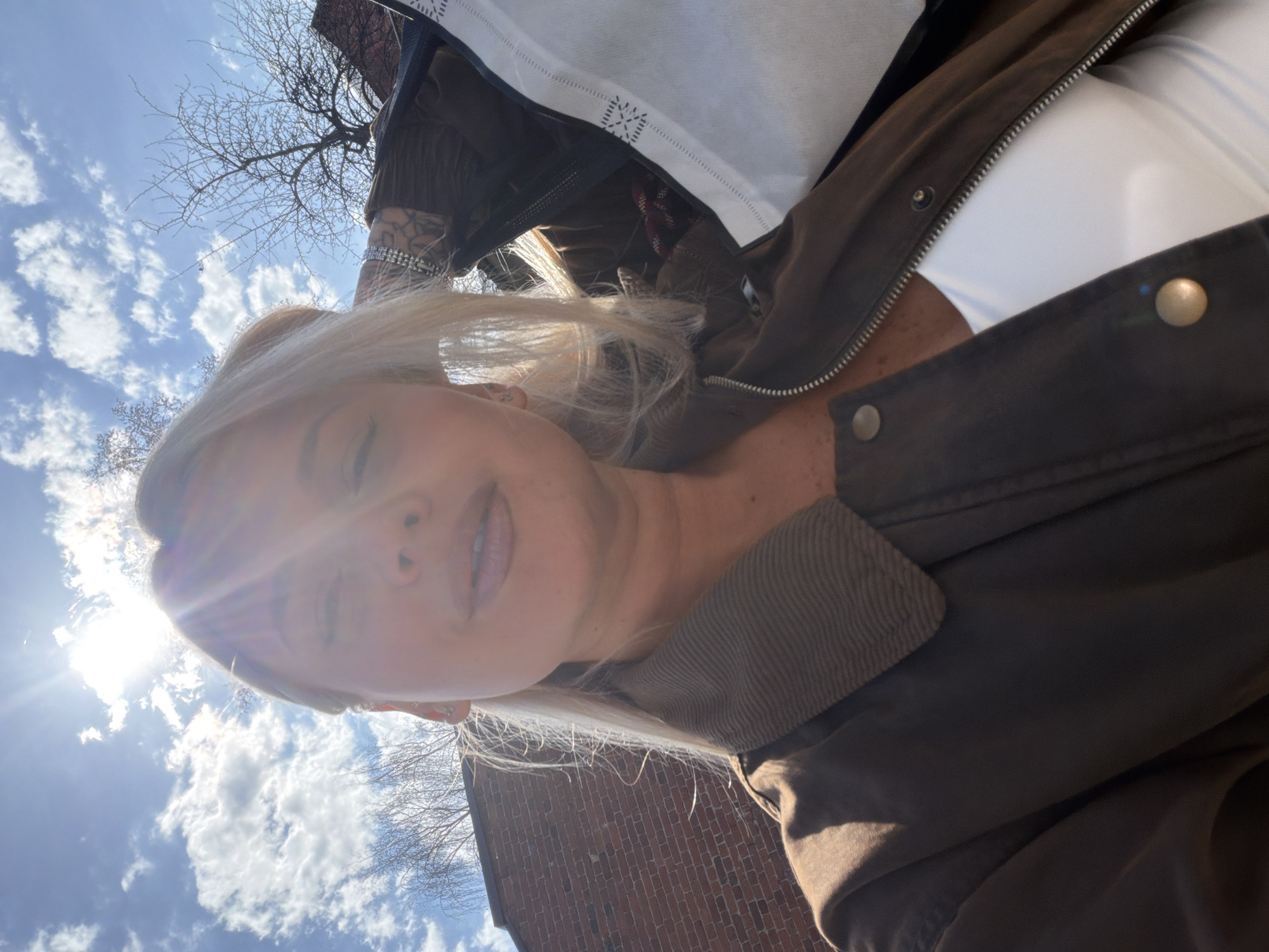 The original image is a low-angle, upward-facing selfie of a woman, presented rotated 90 degrees counter-clockwise. The woman, with blonde hair and fair skin, is visible from the chest upwards. Her face is partially obscured by a bright sun flare originating from the upper left, causing a radiant glow. Her lips are slightly parted, and her expression is neutral.
She is wearing a dark brown or olive-green jacket with a prominent collar, visible zipper, and snap buttons. A light-colored garment, possibly white, is visible underneath her jacket. On her right arm, a multi-layered, sparkling bracelet is worn, and a distinct tattoo pattern can be seen on her forearm, extending up from her wrist.
The background primarily consists of a bright blue sky with scattered white cumulus clouds. The sun is a dominant feature in the upper left, casting strong light and lens flare across the scene. Bare tree branches, characteristic of late autumn, winter, or early spring, are visible against the sky, particularly in the central and right areas. A brick building, indicating an urban or suburban setting, is partially visible in the right background.
The activity depicted is a self-portrait taken outdoors during daytime, likely on a clear, sunny day. No visible text is present in the image.