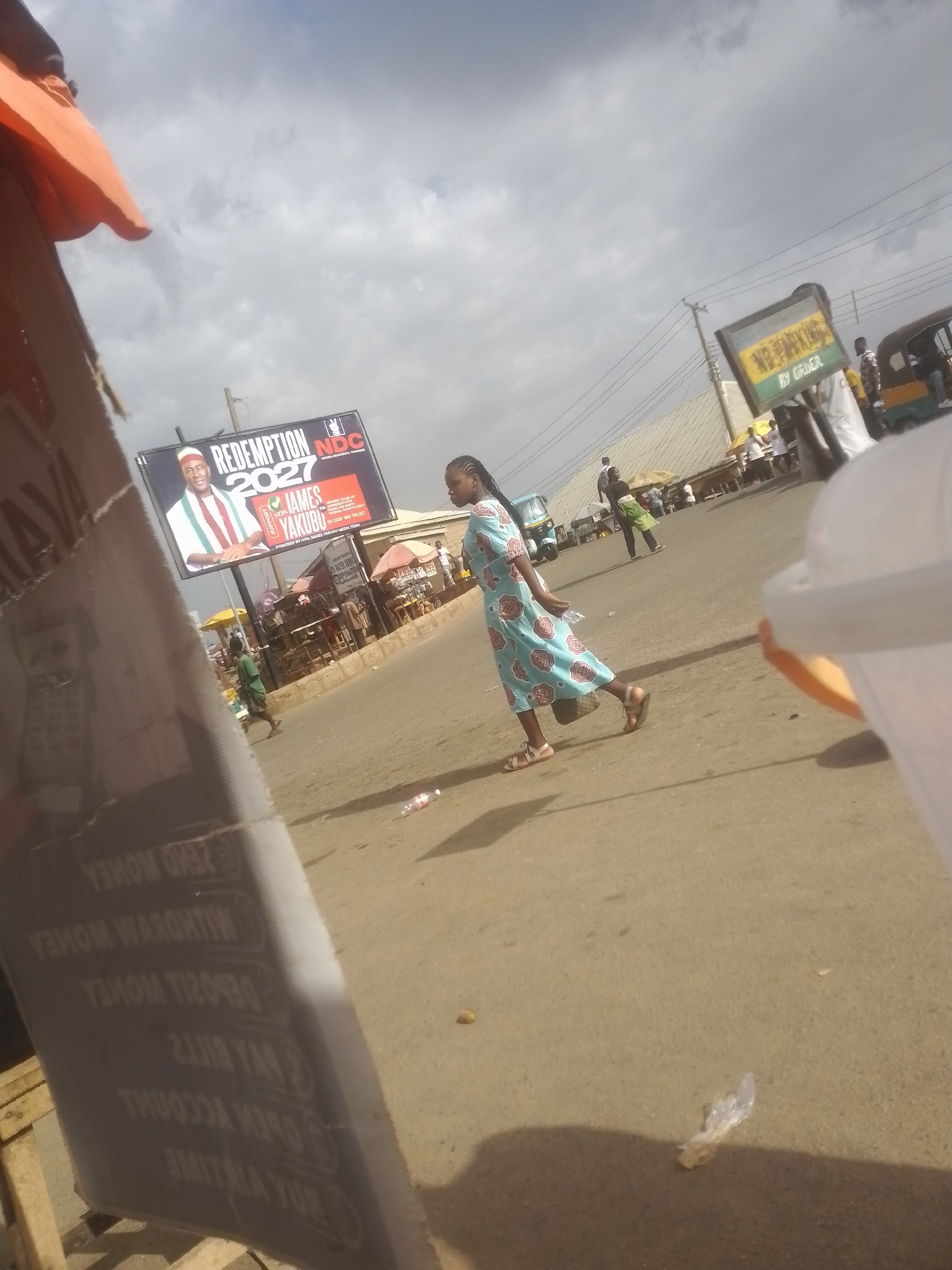 This image shows a street scene with several people and objects. In the foreground, a woman in a light blue and pink patterned dress is walking across a paved road. She is wearing sandals and carrying a dark bag. To the left of the woman, a large, partially obscured sign made of wood and possibly cardboard is leaning against something. The sign has text printed on it, some of which is reversed and appears to be related to financial transactions, such as "SEND MONEY" and "DEPOSIT MONEY".
In the background, a large billboard is visible, featuring a portrait of a man in a white and red striped outfit, with the text "REDEMPTION 2027" and "HON. JAMES YAKUBU". The billboard also includes the logo of "NDC". Further in the background, there are other people, some standing and some walking, as well as small structures that might be market stalls or shops. A yellow and green "NO PARKING BY ORDER" sign is also visible. A three-wheeled vehicle, possibly an auto-rickshaw, is parked on the right side of the image.
The sky is overcast with clouds, suggesting it might be daytime with the sun partially obscured. The overall impression is of a busy street in a populated area, possibly a market or commercial district. There are some scattered pieces of trash on the road, including a plastic bottle and some crumpled plastic.