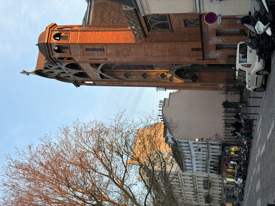 The image depicts a street scene in the 19th arrondissement of Paris, France, specifically featuring the Église Saint-Jean-Baptiste de Belleville. The large Gothic Revival church, constructed of red brick, dominates the right side of the frame. Its ornate bell tower features intricate tracery, arched openings, and a cross-topped spire, all illuminated by warm, low-angle sunlight. The church's lower facade displays tall, narrow windows and a decorative street lamp integrated into the entrance archway; graffiti is visible on the lower brickwork to the right.
To the left, a large, bare deciduous tree with branches catching the sunlight partially obscures several multi-story Haussmannian-style buildings with light-colored facades and balconies. Further back, a plainer building with a beige facade stands adjacent to the church, and a distant building with a rounded roof is visible on the horizon, also bathed in warm light.
The street is paved with cobblestones, and numerous scooters and motorcycles are parked in rows along the left side of the thoroughfare. A white compact SUV is parked in the foreground on the right, adjacent to a street sign indicating "No Parking Except" ("SAUF"). Several pedestrians are visible on the sidewalks, including individuals near the parked vehicles and on the left side of the street. Black bollards line the sidewalk. The sky is clear blue, and the overall lighting suggests either late afternoon or early morning.