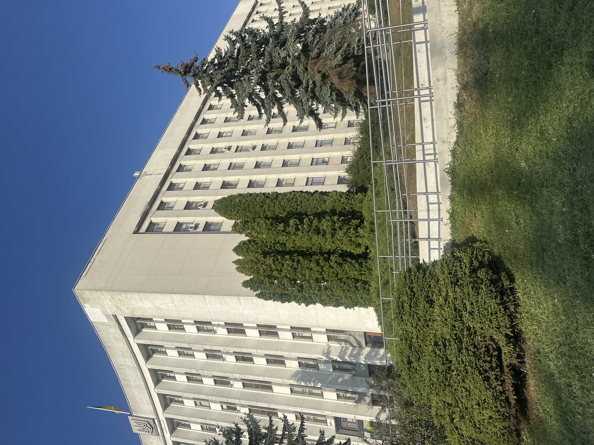 The image shows the Ternopil Regional State Administration building, a large, concrete, multi-story structure with a symmetrical facade featuring numerous windows. The building is adorned with the Ukrainian coat of arms and the national flag flying from the rooftop. In front of the building, a well-maintained landscape includes manicured bushes, tall evergreen trees, and a grassy lawn. A metal railing runs along a pathway in front of the bushes. The scene is bathed in the bright light of a sunny day, casting sharp shadows on the building's facade and the surrounding grounds. The sky is a clear, deep blue, suggesting fair weather.