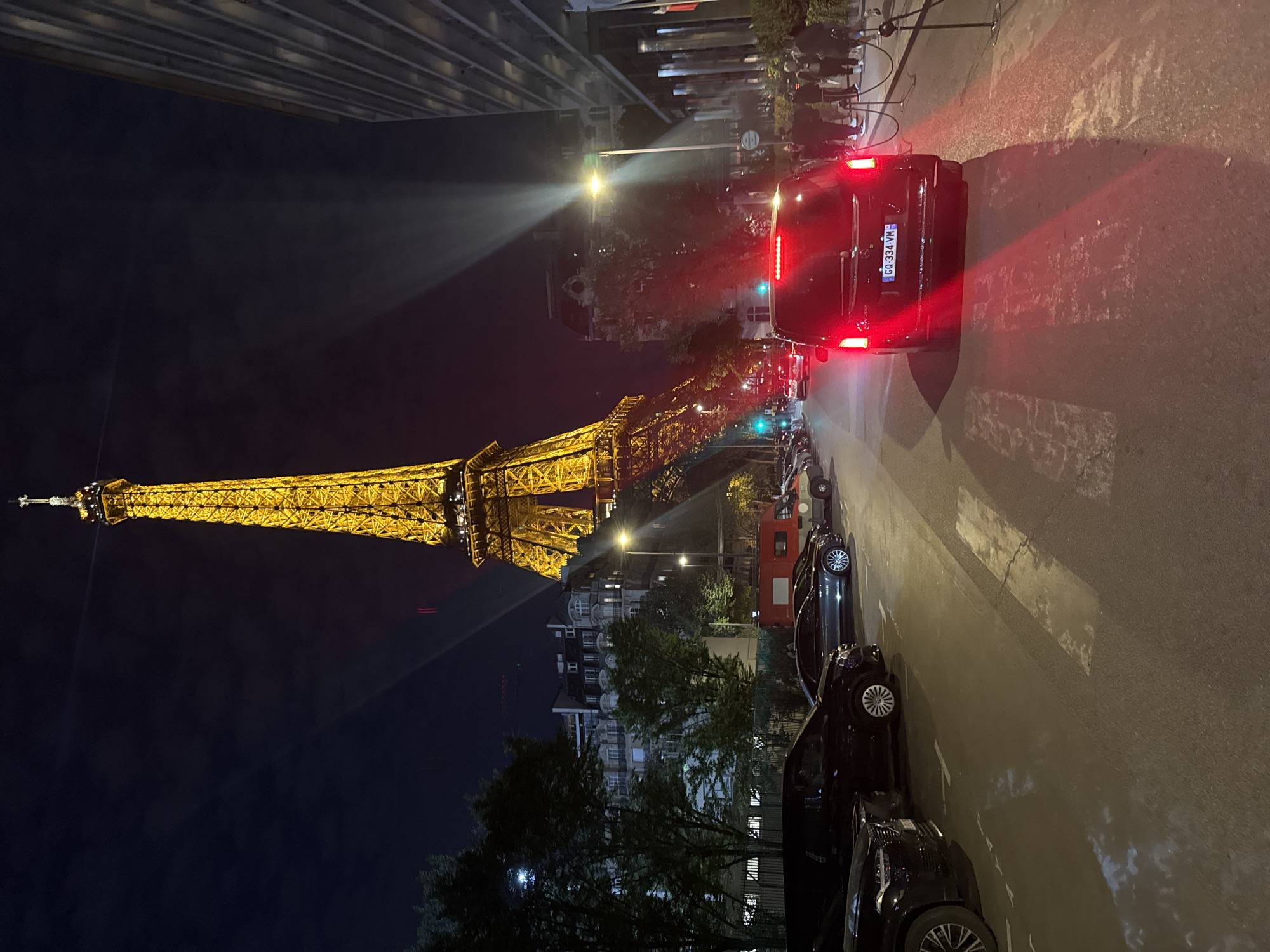 This is a night view in Paris, France, featuring the illuminated Eiffel Tower in the background. A street runs alongside the tower, lined with parked cars on the right. A black van with its taillights glowing is stopped in the middle of the street. People are standing on the sidewalk to the right. Buildings and trees are visible along the street. A beam of light stretches across the scene from a street lamp. The sky is dark.