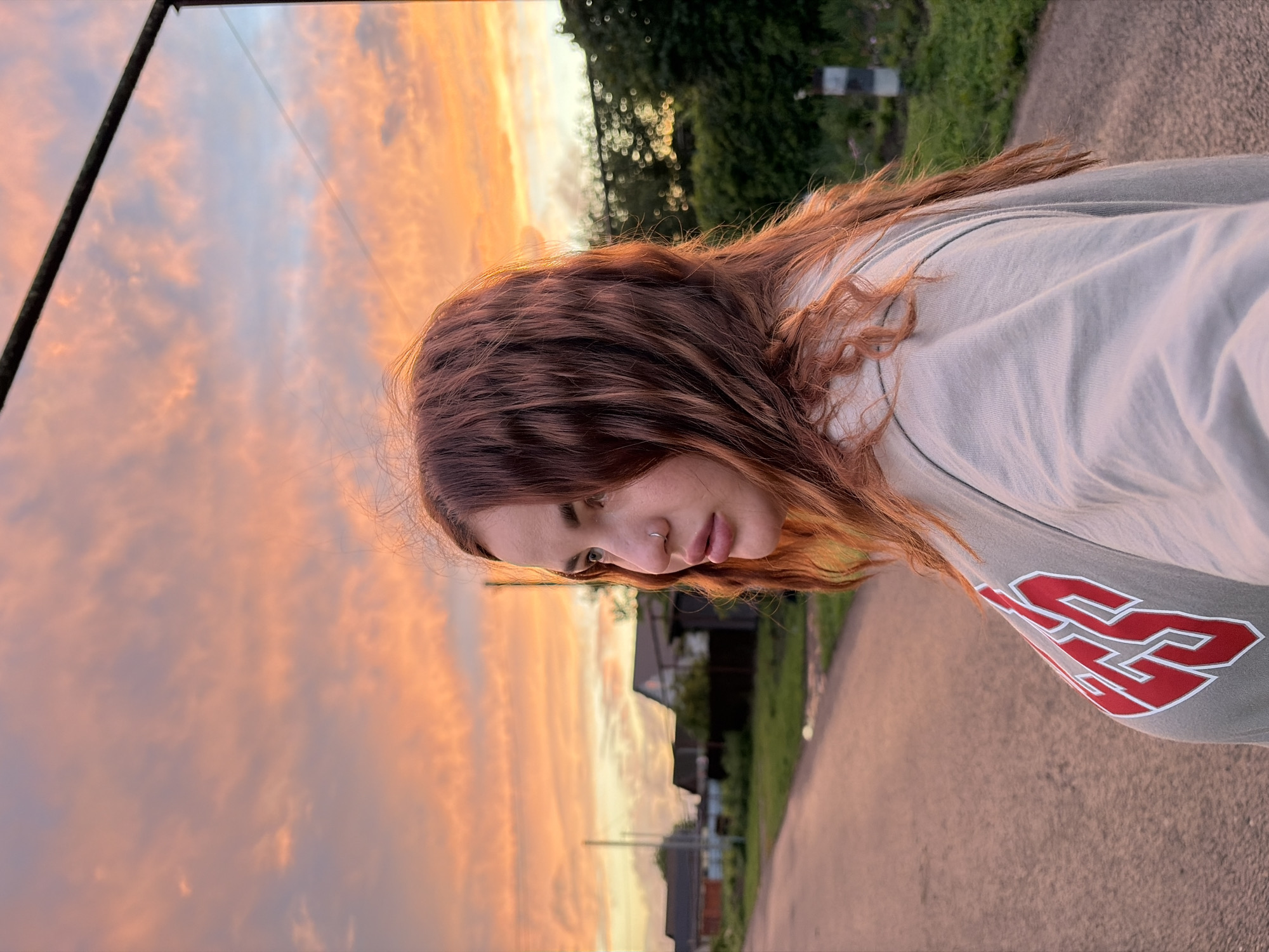 The image shows a young woman standing on a paved road in an urban or suburban setting, possibly in Unknown, Unknown. Her hair is reddish-brown, and she is wearing a light gray or green sweatshirt with a red and white design. She is looking down. In the background, there are houses and trees. The sky is a vibrant orange and pink, suggesting it is either sunrise or sunset. The image appears to be taken in a portrait orientation. The left side of the frame features a reflection of the sky.