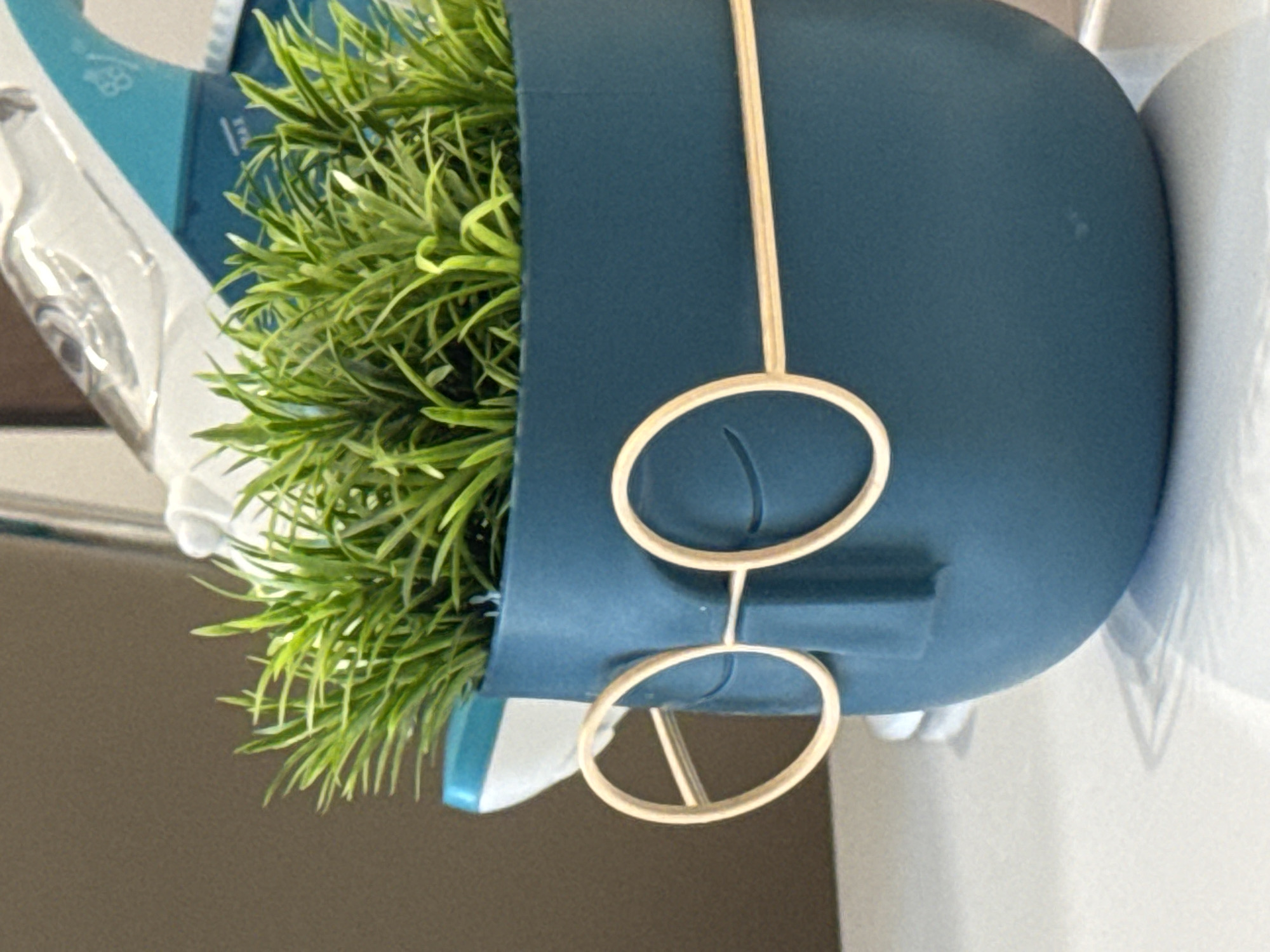 The image shows a close-up of a teal-colored ceramic planter shaped like a face with a minimalist design. The planter has two round, gold-colored wire glasses perched on its "nose" area, and two thin, curved lines representing closed eyes. Inside the planter, there is a cluster of vibrant green artificial grass. In the background, out of focus, is a portion of a white and teal steam iron. The planter sits on a light-colored, reflective surface, likely a tabletop or shelf. The lighting suggests an indoor environment. The image does not contain any information or visual cues to determine its location in Plovdiv, Bulgaria.