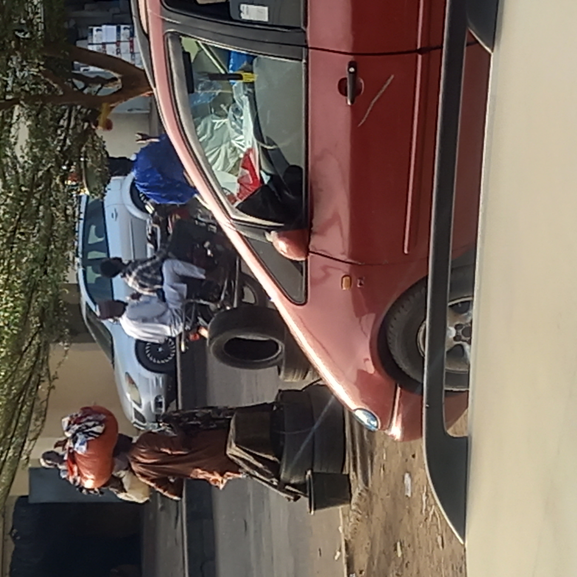 This image captures a vibrant street scene, likely in Dadi Riba, Nigeria, under clear daylight conditions. The ground appears dusty and unpaved in many areas, characteristic of local roads.
In the foreground, a person wearing a brown garment is seen carrying a large, bright orange bundle, possibly filled with goods or textiles, balanced on their head. They are positioned near several stacked car tires on the ground.
A prominent red car, possibly a minivan or SUV, occupies the midground, parked sideways relative to the viewer. Its interior, visible through the window, appears filled with various items. A distinct scratch mark is visible near the car's door handle.
Further back, a white car is parked alongside. Gathered near this white car and a motorcycle are several individuals. Two men are clearly visible: one wearing a plaid shirt and light-colored trousers, and another in a lighter-toned shirt. A third person is partially seen seated on the motorcycle, which also has a spare tire attached. Lush green foliage from trees lines the left side of the frame, suggesting some natural shading.
The scene conveys everyday activity, with people going about their routines, possibly engaging in commerce or travel. No discernible text is visible within the image.