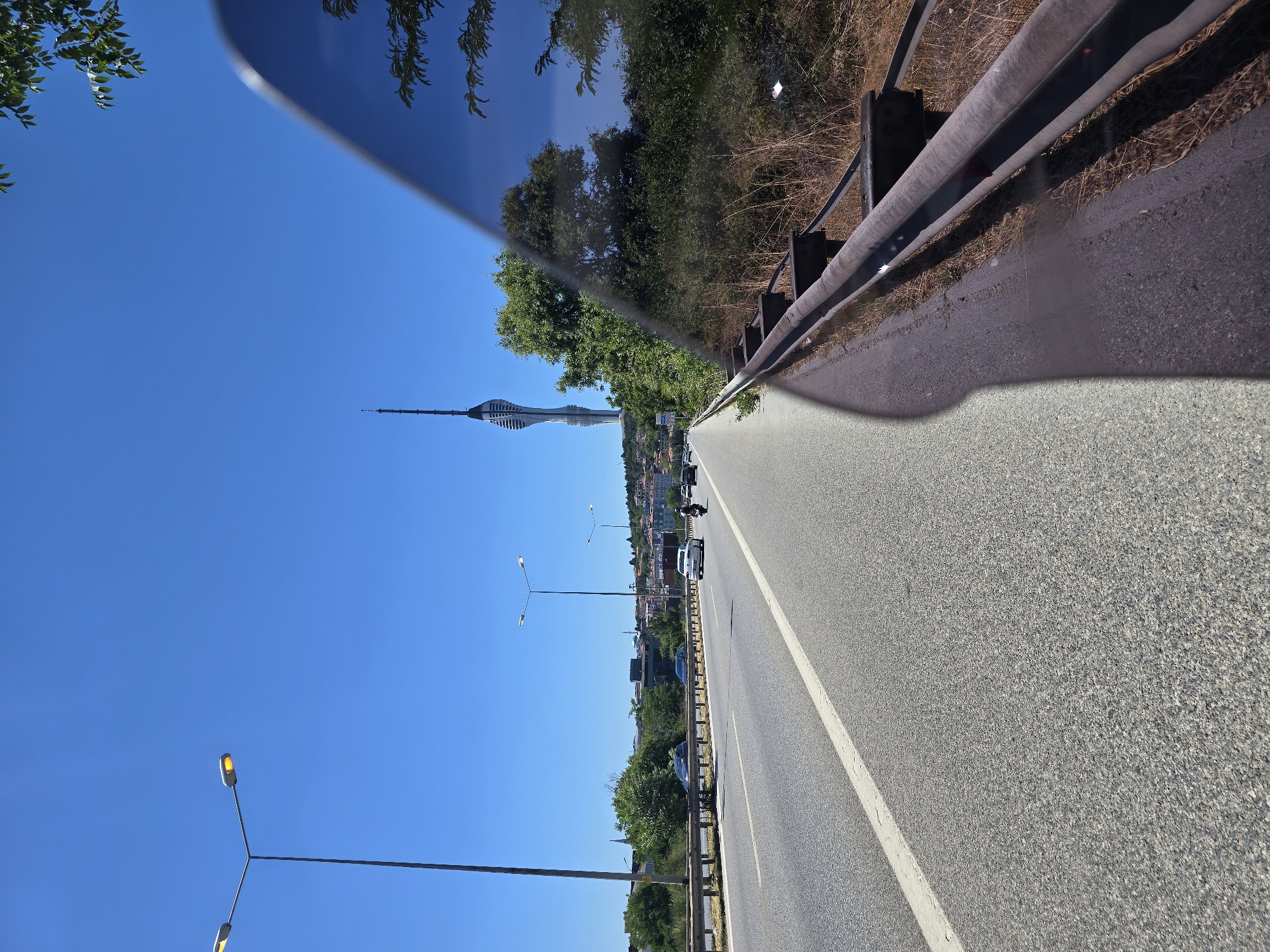 The image captures a daytime view of a highway in Üsküdar, Turkey, with the Çamlıca Tower prominently visible in the distance. The highway has multiple lanes with traffic, including cars and a motorcycle. Streetlights line the road. The right side of the image shows a metal guardrail and dense foliage, with reflections visible on a dark surface, possibly a vehicle's side mirror or window. The sky is clear and blue.