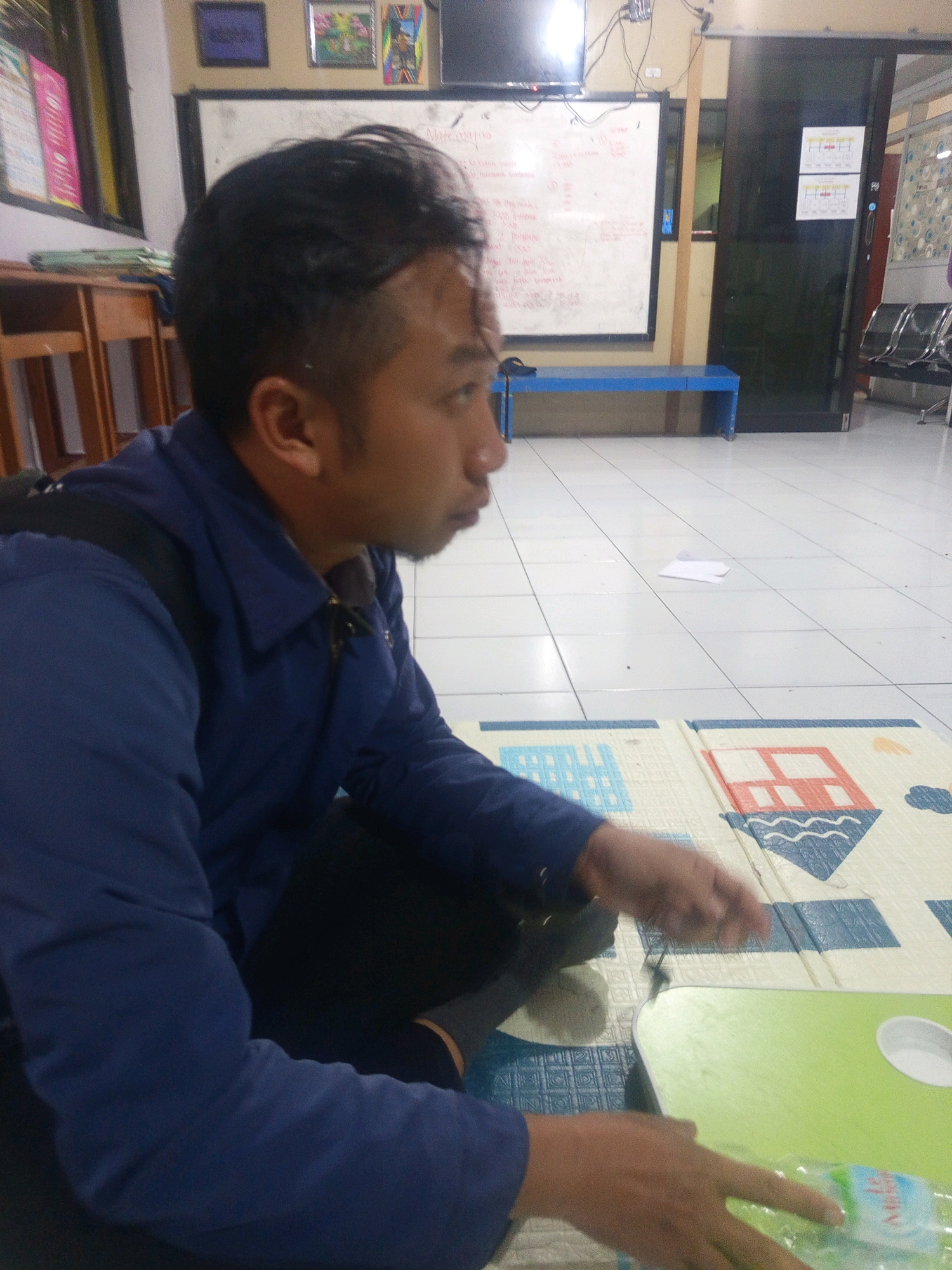 A medium shot captures a man with short black hair and a light brown complexion, facing right in profile. He is wearing a dark blue collared shirt, with a black backpack strap visible over his left shoulder. He is seated on a floor, leaning slightly forward, with his hands positioned in front of him. His right hand is holding a clear plastic water bottle with a blue and white label.
The man is in a room with a tiled floor, laid out in a grid pattern. To his right, on the floor, is a large, colorful play mat with a cartoon house design. In the background, there is a whiteboard with writing on it, and a blue bench positioned below it. To the left, a wooden shelf unit is visible. Further in the background, a dark sliding door and a doorway with chairs can be seen. The scene appears to be an indoor public space, possibly a classroom or community area, with elements suggesting a casual or educational setting.