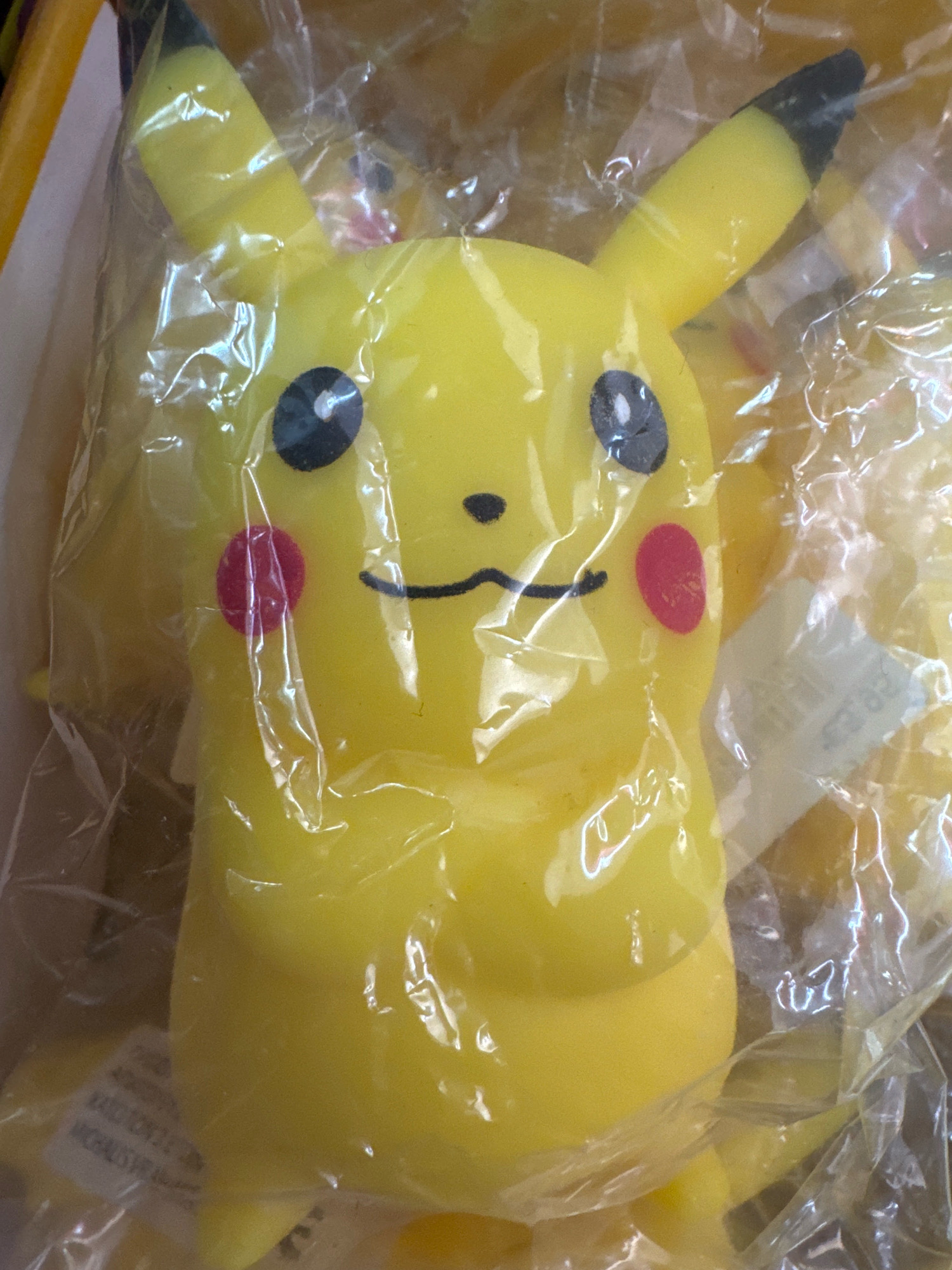 The image shows a yellow Pikachu toy, likely made of plastic or rubber, enclosed in clear plastic packaging. The toy is depicted from the chest up, with its characteristic pointed ears, black eyes with white pupils, and red circular cheeks visible. A black curved line forms its mouth. The toy is illuminated from the front, casting subtle shadows and highlights on its surface and within the packaging. In the lower left corner of the image, some text on a label is partially visible, appearing to be English writing. The background is out of focus and appears to be a light-colored surface. There are no people or animals present in the image, and no environmental context is provided beyond the packaging. The image does not contain any information related to the city of Limassol, Cyprus.