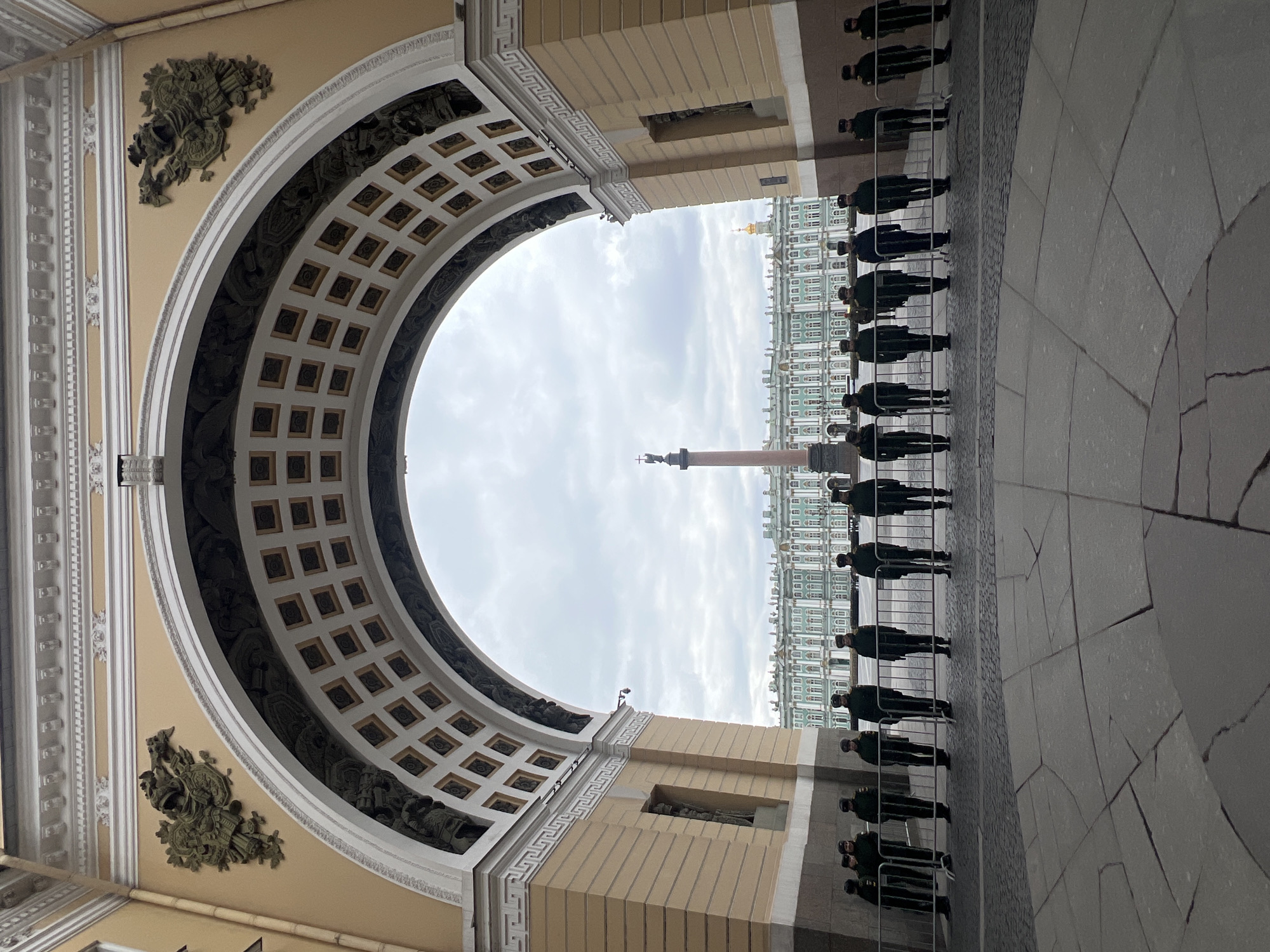 This image captures a view from beneath a grand, ornate archway in Saint Petersburg, Russia, likely part of the General Staff Building framing Palace Square. The arch, made of light-colored stone, features a deeply coffered ceiling with square panels and intricate sculptural reliefs on its inner surface and spandrels, showcasing classical architectural details.
Beyond the arch, an expansive paved square is visible. In the distance, a tall, slender column (the Alexander Column) with a statue on top stands prominently. Behind the column, the magnificent facade of a large, multi-story building (the Winter Palace/Hermitage Museum) stretches across the horizon. This distant building is characterized by its light green and white color scheme, numerous windows, and elaborate architectural ornamentation, with a golden dome visible above its roofline.
Along the right side of the paved square, a line of approximately ten uniformed individuals, possibly guards or military personnel, stand in formation behind black metal barriers. They are dressed in dark uniforms and caps, facing the distant palace. The ground consists of large, light-colored flagstones transitioning to cobblestones closer to the line of people.
The sky is overcast with soft grey clouds, suggesting a daytime scene without direct sunlight. The atmosphere conveys a sense of historical grandeur and formal observance in this iconic Russian location. No visible text is present in the image.