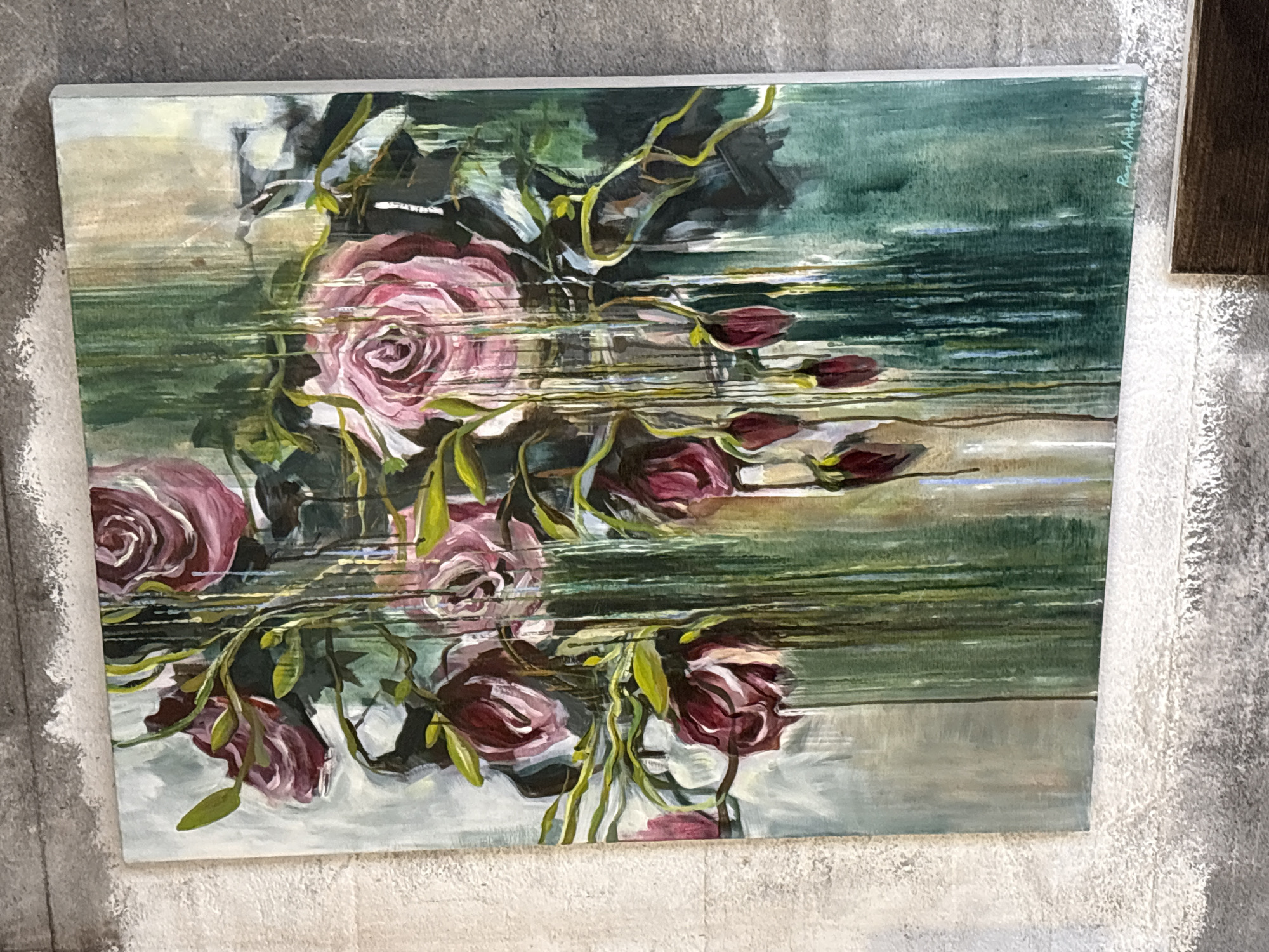A vertical, framed oil painting of a bouquet of pink and deep red roses with green leaves and stems hangs on a textured, light-colored wall, possibly concrete, that has a rough finish. The painting depicts a somewhat abstract arrangement of roses, with some fully bloomed and others in bud form. Vertical streaks of green, gold, and blue paint are layered over and behind the roses, giving the impression of rain or dripping water. The background of the painting is a blend of muted greens, grays, and hints of beige. The bottom right corner of the painting has a faint, illegible signature. Below the painting, a dark brown wooden edge of a piece of furniture is visible. The overall impression is of a moody floral still life. The specific location in Gryllefjorden, Norway, is not ascertainable from the image.