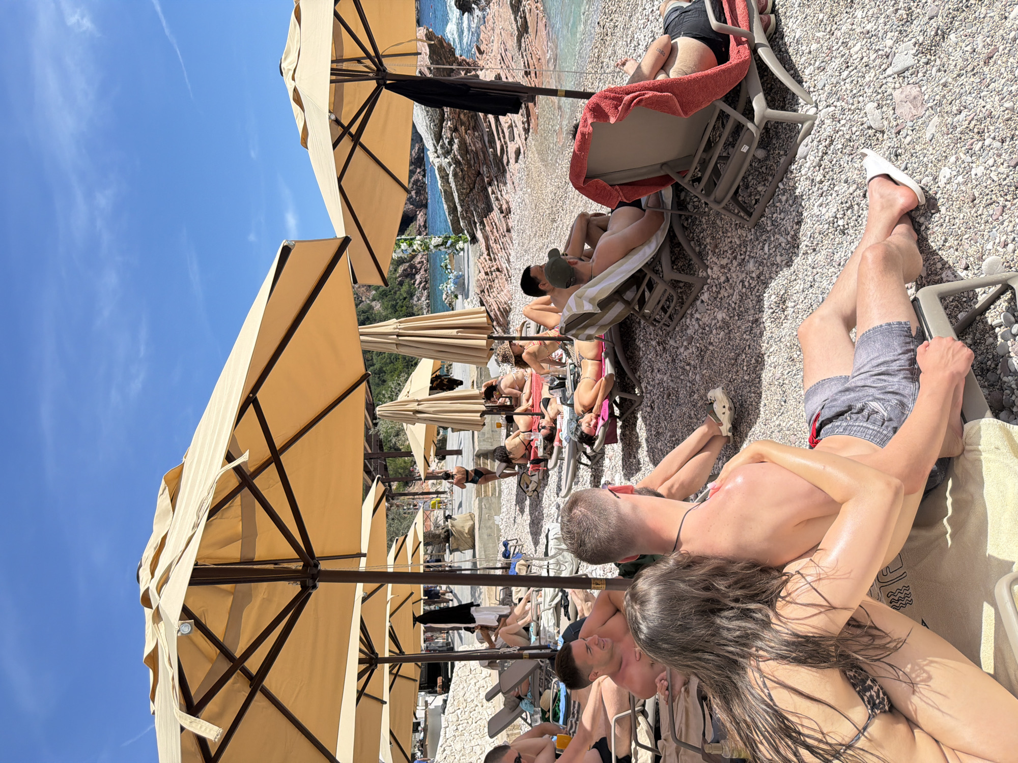 A beach scene in Sveti Stefan, Montenegro, shows multiple sunbathers on lounge chairs under a row of tan umbrellas. The beach is covered in light-colored pebbles. In the foreground, a couple is visible, with a woman in a patterned bikini and a man in gray shorts. Further back, other individuals are relaxing on chairs, some reading or lounging. The background features rocky cliffs and turquoise water.