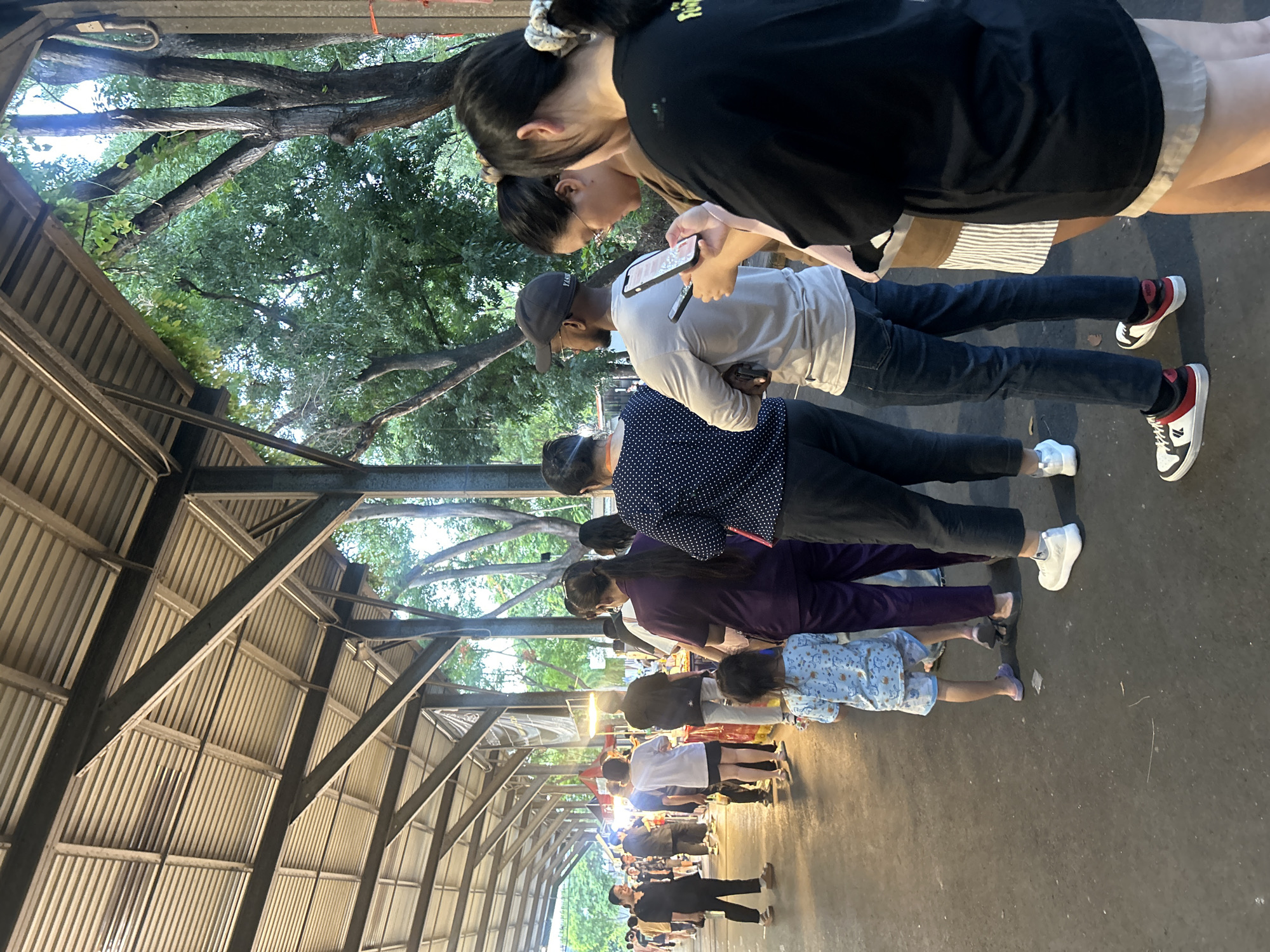 The image depicts a street scene at dusk in Tha Khlong Town Municipality, Thailand. A covered walkway with a corrugated metal roof supported by metal beams stretches into the distance, illuminating a food stall area with overhead lights. A line of people, varying in age and gender, are queuing. In the foreground, a woman in a black shirt and shorts faces away from the camera, while to her right, a young girl in a light blue patterned outfit stands with her back to the viewer. Further back, several individuals are visible in the queue, some looking at their phones. The ground is paved with asphalt. Trees with green foliage are visible behind the walkway, suggesting an outdoor market setting. The overall atmosphere appears casual and public, likely a common gathering place for food or shopping.