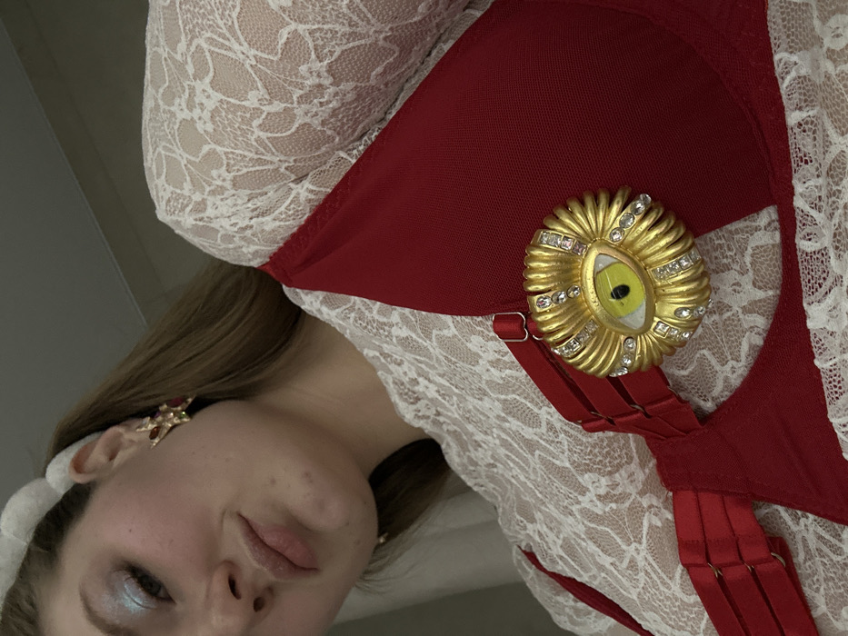 A woman with fair skin and light brown hair is visible from the shoulders up. She wears a white lace top under a red garment resembling a bra or corset with several straps. A gold brooch shaped like an eye, adorned with small gems, is pinned to the red fabric. Her makeup includes light blue eyeshadow. A decorative earring is visible on her left ear.