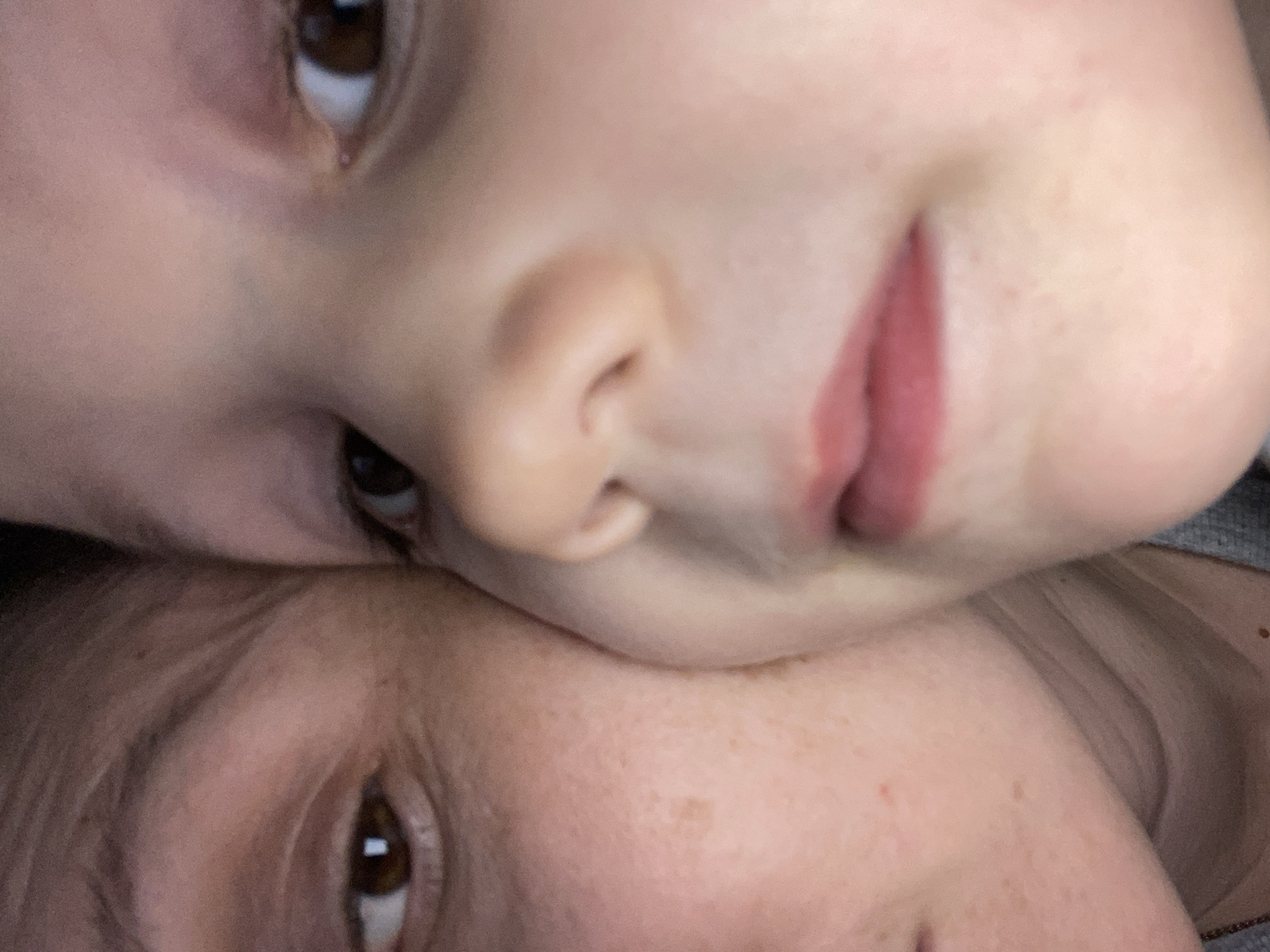 The image shows a close-up view of two individuals. The top individual is a young child with fair skin, brown eyes, a small nose, and pink lips. The bottom individual is another person, also with fair skin and brown eyes. The image composition shows the faces closely positioned together, with the child's face tilted at an angle. No information is provided regarding the location in the city of Noviy, Russia.