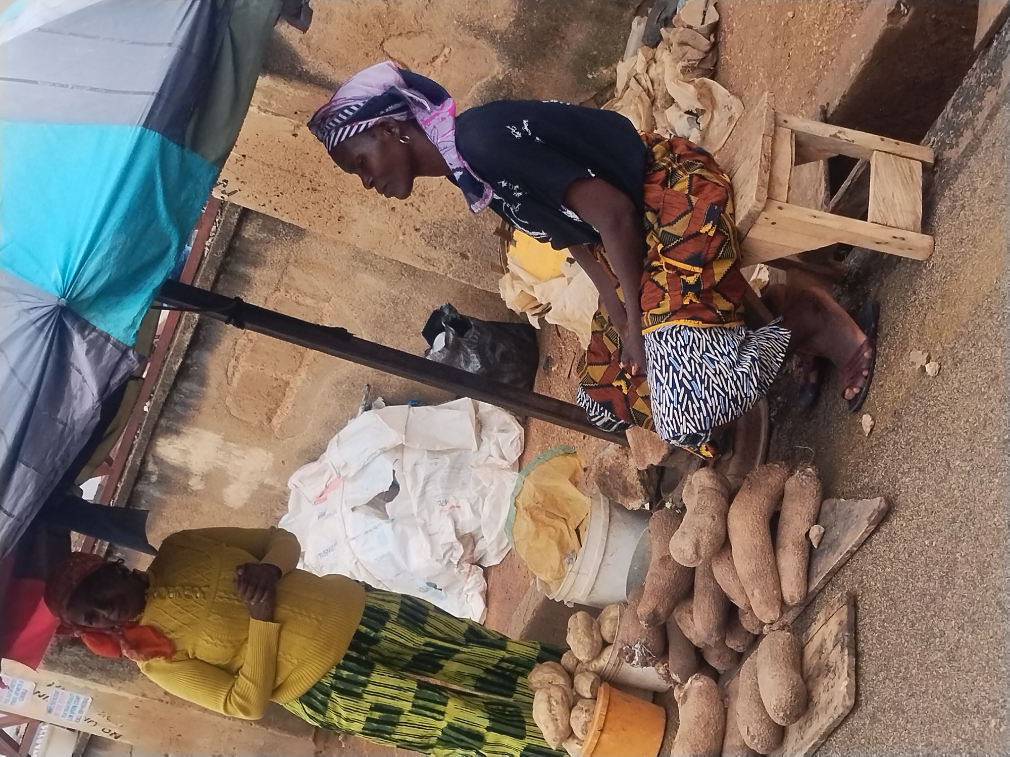 Two women are positioned outdoors next to an array of goods laid out on the ground. One woman is standing behind a small wooden stool, leaning forward slightly as if examining the items laid out before her. She's wearing a dark-colored shirt, a patterned skirt, and a headscarf. Her feet are clad in sandals. The other woman is standing with her arms folded across her chest, wearing a yellow sweater and a green-striped skirt with a red headscarf. To their sides are piles of what appear to be root vegetables, possibly yams and potatoes, as well as containers and bags. A blue and grey umbrella is partially visible, offering shade over the scene. The setting appears to be an outdoor market area.
