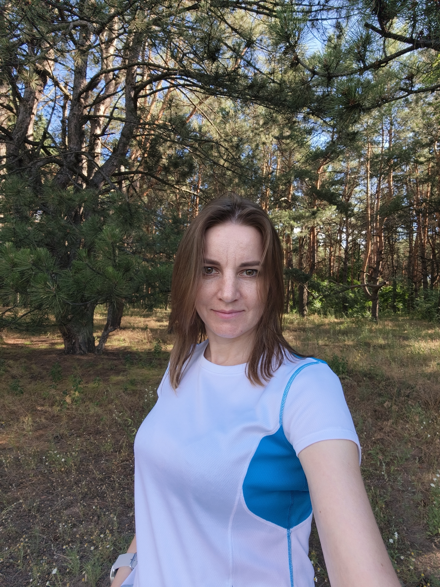 A woman is taking a selfie in a forest. She is wearing a white t-shirt with blue accents and a smartwatch on her left wrist. She has brown hair and is looking at the camera with a slight smile. The background is a forest with tall pine trees and dry grass on the ground. The sun is shining, indicating it is daytime, and the sky is blue and clear. The setting appears to be a nature trail or a park. The woman seems relaxed and enjoys the outdoors. The location is Shyroke, Ukraine. There is no visible text in the image.