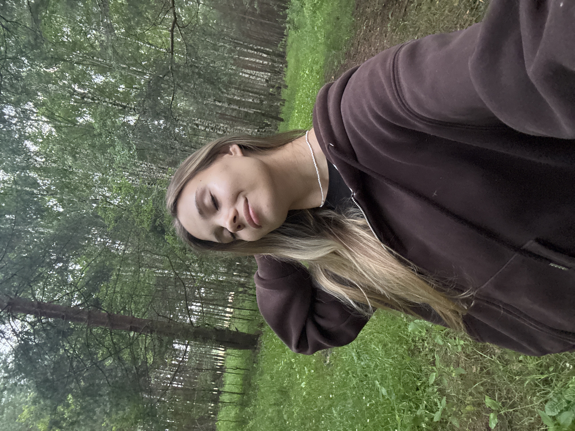 The image shows a young woman with blonde hair, wearing a dark brown hooded jacket and a silver necklace. She is posing for a selfie, with her eyes closed and a slight smile on her face, conveying a sense of peace or contentment. The background is a lush green forest with tall trees and dappled sunlight filtering through the canopy, suggesting it is daytime and the weather is pleasant. The woman is positioned in the foreground, with the trees and grass filling the rest of the frame. The orientation of the image is rotated, with the woman appearing on the left side and the forest on the right. The overall atmosphere is serene and natural, hinting at an outdoor excursion. The image has been cropped with a vertical orientation.