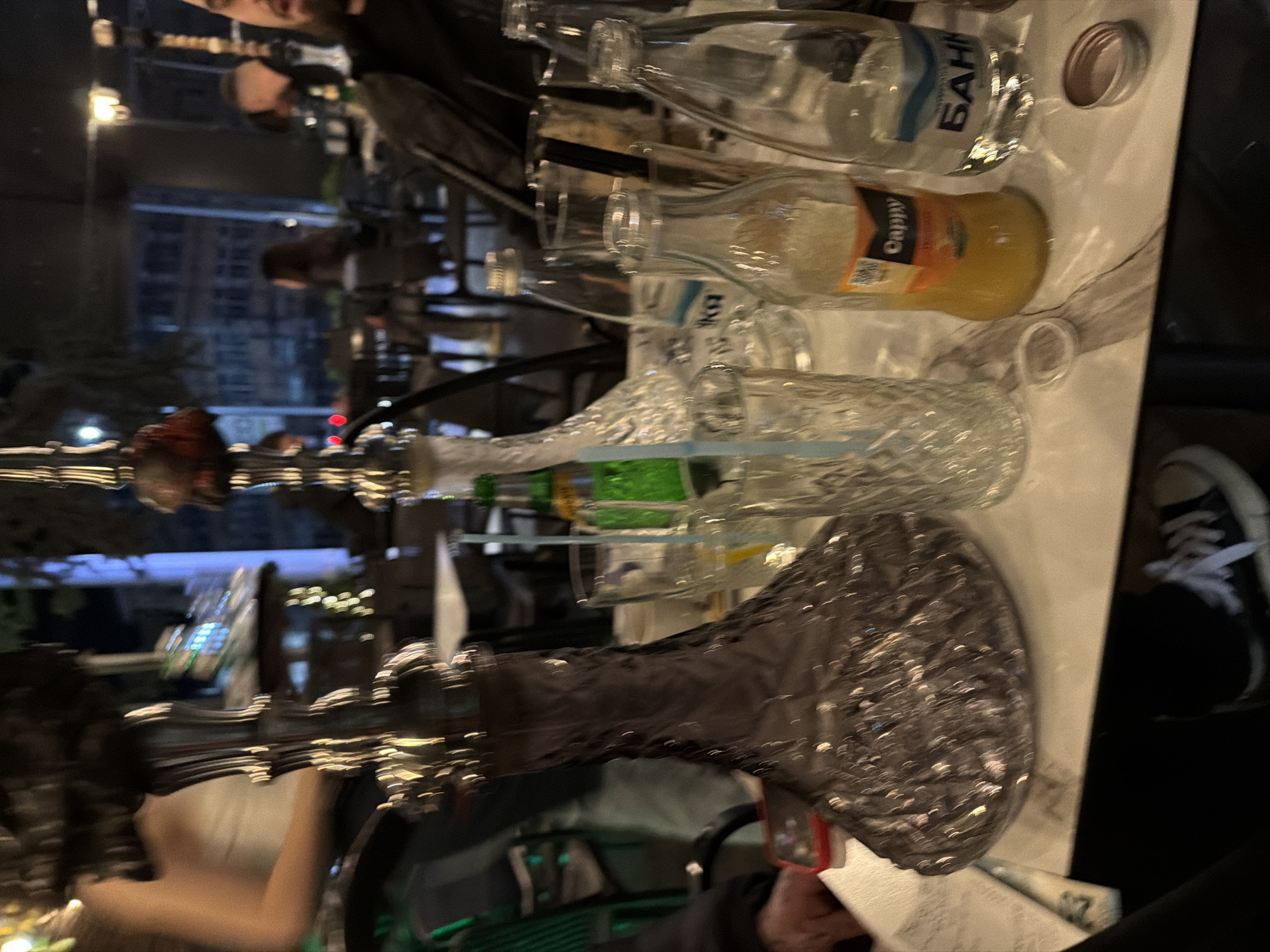 The image captures a table scene, presumably at a café or restaurant, located in Unknown, Unknown. The composition features a hookah as the focal point, with its silver-toned base prominently displayed. Several bottles sit on the table, including one labelled "Capry" filled with an orange-colored liquid. A bottle with Cyrillic script is also visible, alongside a metal lid. A glass with a crystalline pattern is also present.
In the background, a blurred cityscape is visible through a window, suggesting an urban setting. People are present in the background. The presence of the hookah, along with beverages, suggest a social gathering or relaxation setting. A pair of black and white sneakers partially enter the frame from the side. A hand holding a red phone is also present. The table itself is light-colored, with a surface suggesting it is made of stone.