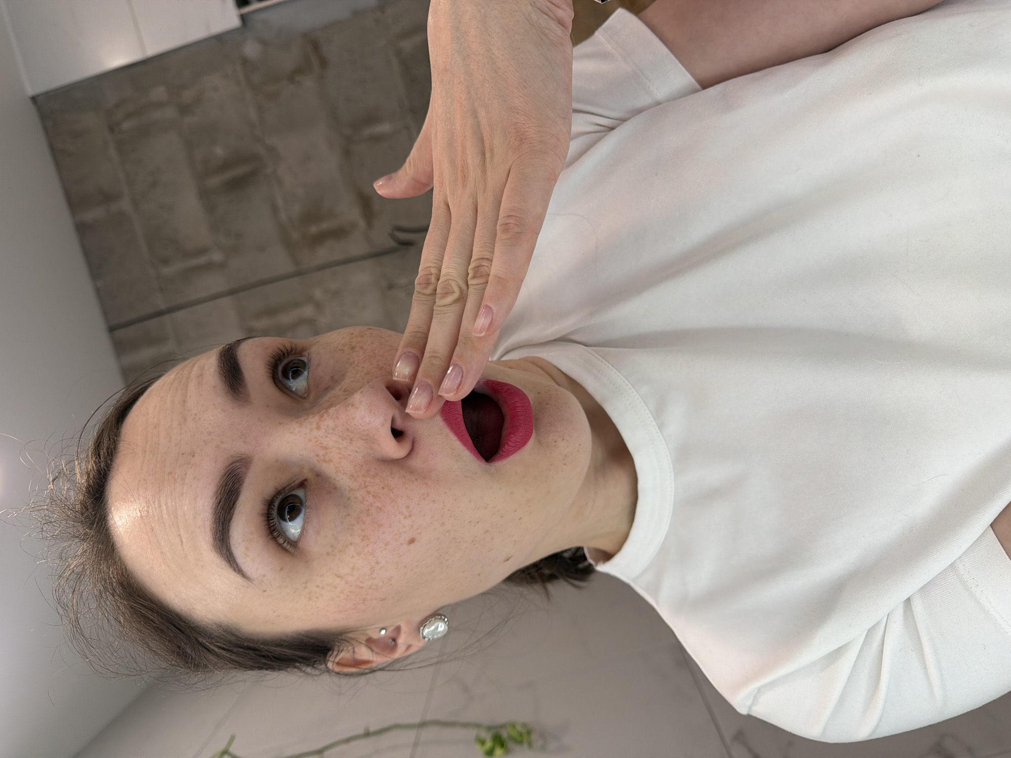 A woman with freckled skin and dark hair, wearing a white T-shirt, is positioned in front of a textured, neutral-toned wall. She has red lipstick and is holding one hand near her mouth, her fingers touching near her lips. A pair of earrings can be seen on her left ear. The background includes a glimpse of flooring and plant life. There is no information to associate this image with Chaikovsky, Russia.