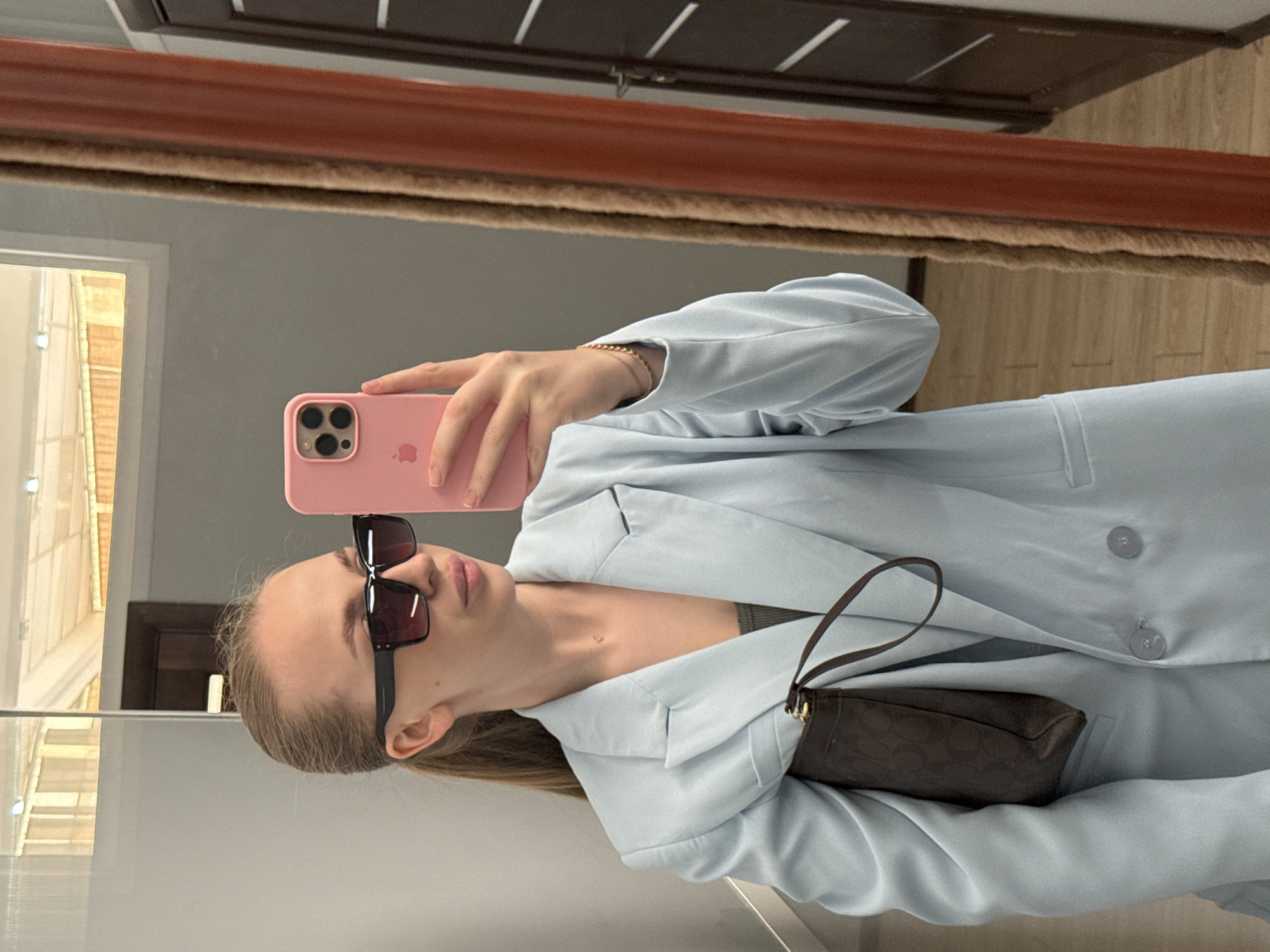 A young woman is depicted from the mid-torso upward, taking a mirror selfie indoors. She has light-colored hair, pulled back into a ponytail. She wears large, dark-framed rectangular sunglasses that obscure her eyes. Her lips are visible and appear naturally colored or lightly glossed. She is wearing a light blue, double-breasted blazer over a dark olive-green or grey tank top. A delicate, small pendant necklace is visible on her chest. Her left arm is holding a dark brown handbag with a subtle patterned texture and a short shoulder strap. Her right hand, adorned with a gold-colored bracelet, holds a pink Apple iPhone with a pink case, positioned to capture the reflection.
The background, visible in the mirror's reflection, shows an interior space. To the left, a light grey wall is visible, with a horizontal windowpane above reflecting an exterior scene. The exterior appears to be a building with light-colored walls and a roofline, suggesting an urban or suburban environment during daylight hours. To the right of the woman, a textured, light brown curtain or drapery is reflected vertically, adjacent to a dark brown door featuring multiple horizontal white or light-colored panels and a visible doorknob. The floor in the reflection is light-colored, possibly wood or laminate. The ceiling, also reflected, is white and features recessed lighting. The specific location within the city of Petropavlovsk-Kamchatsky, Russia, cannot be definitively determined from the visual elements present in the image.