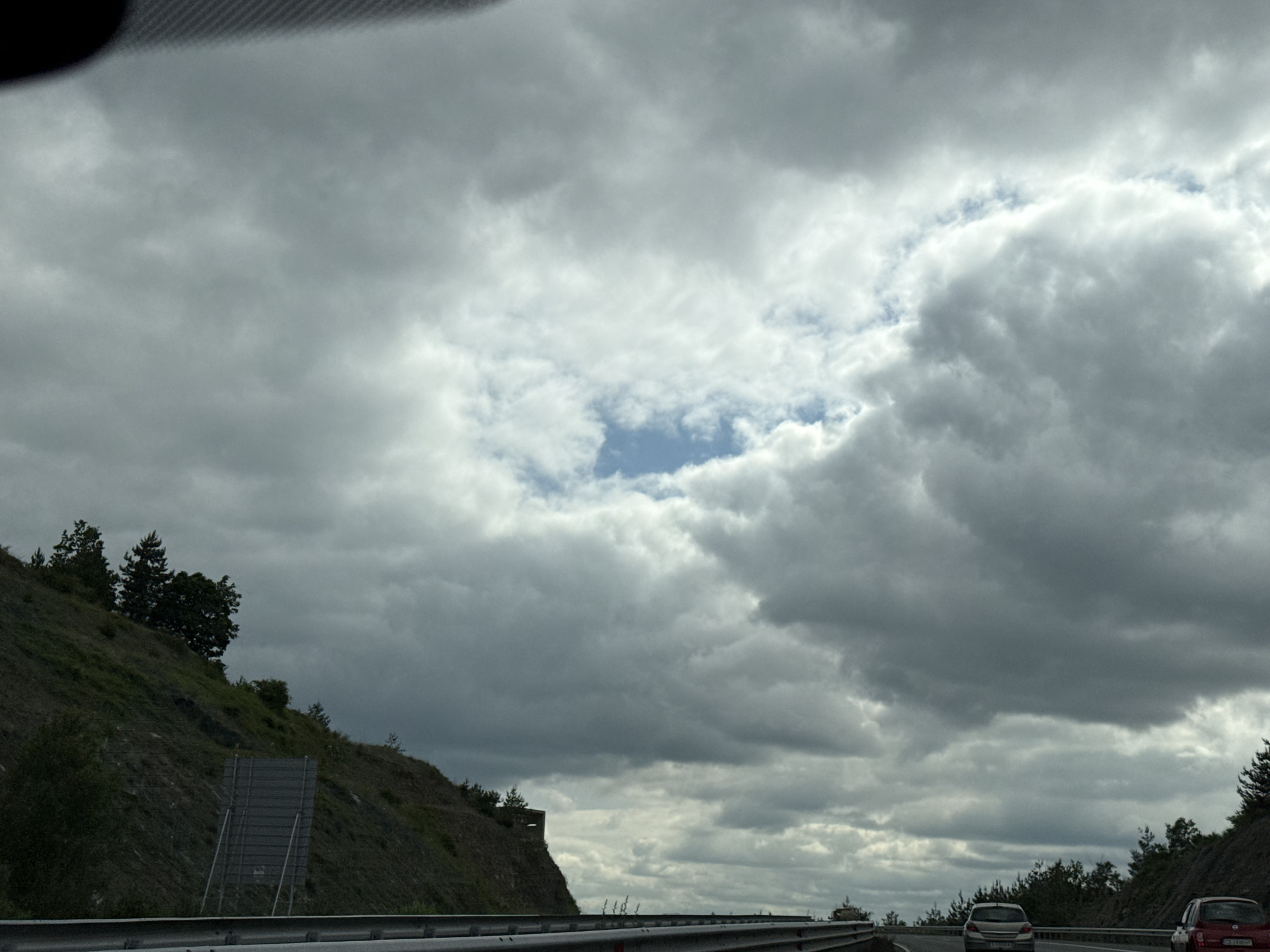 This is a landscape photograph taken from a moving vehicle on a highway. The sky is overcast with heavy, dark clouds, but there is a patch of blue sky visible in the upper center. To the left, a steep, grassy hillside is covered with trees, and a metal guardrail runs along the bottom of the image. Two cars are visible on the highway ahead, suggesting the vehicle the photo is taken from is also on the highway. The scene appears to be on the outskirts of Staro Selo, Bulgaria, given the characteristic landscape of the region. The overall mood of the image is somewhat somber due to the cloudy sky.